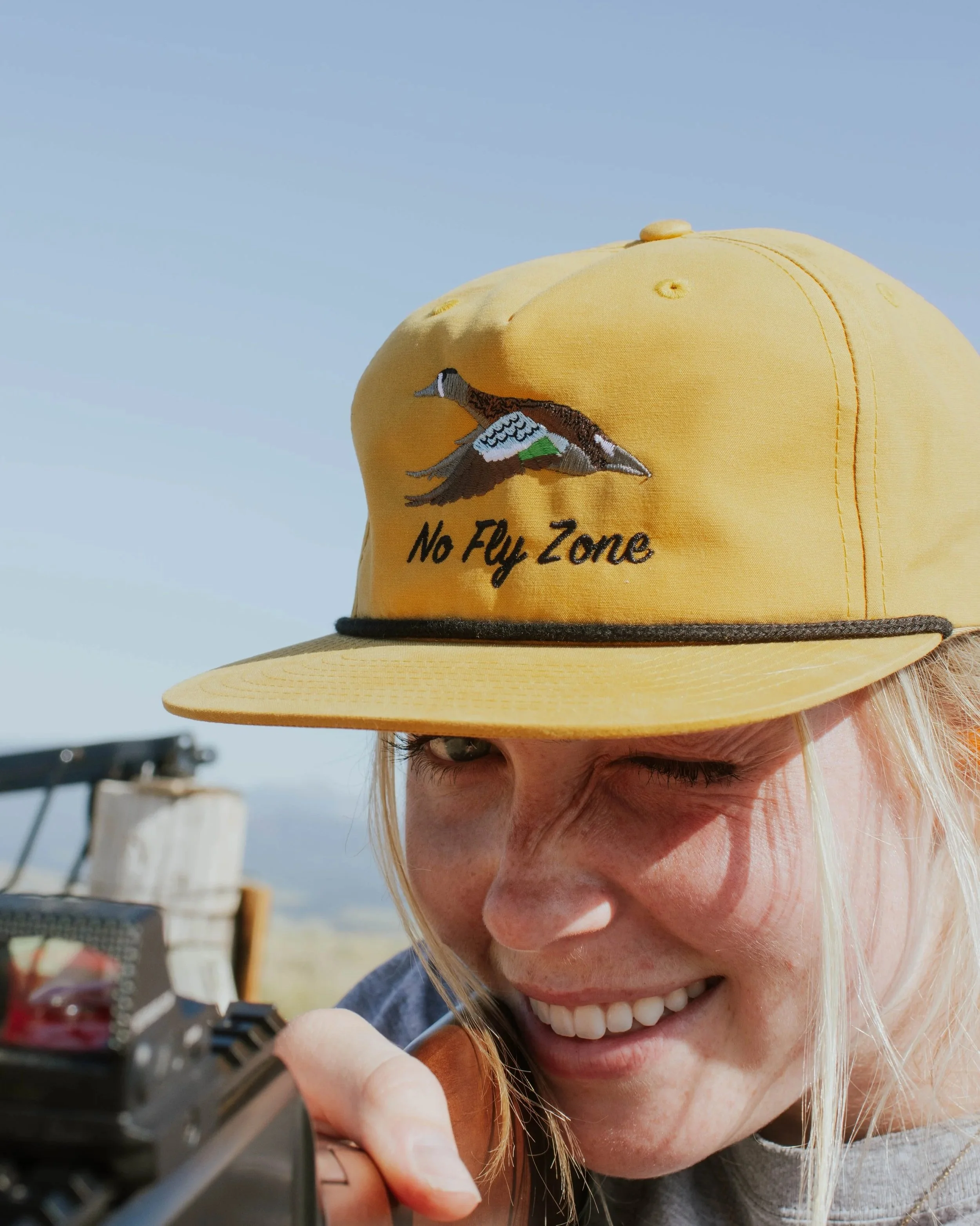 Person aiming a rifle in outdoor shooting range, wearing a yellow cap with a duck embroidered and 'No Fly Zone' text.