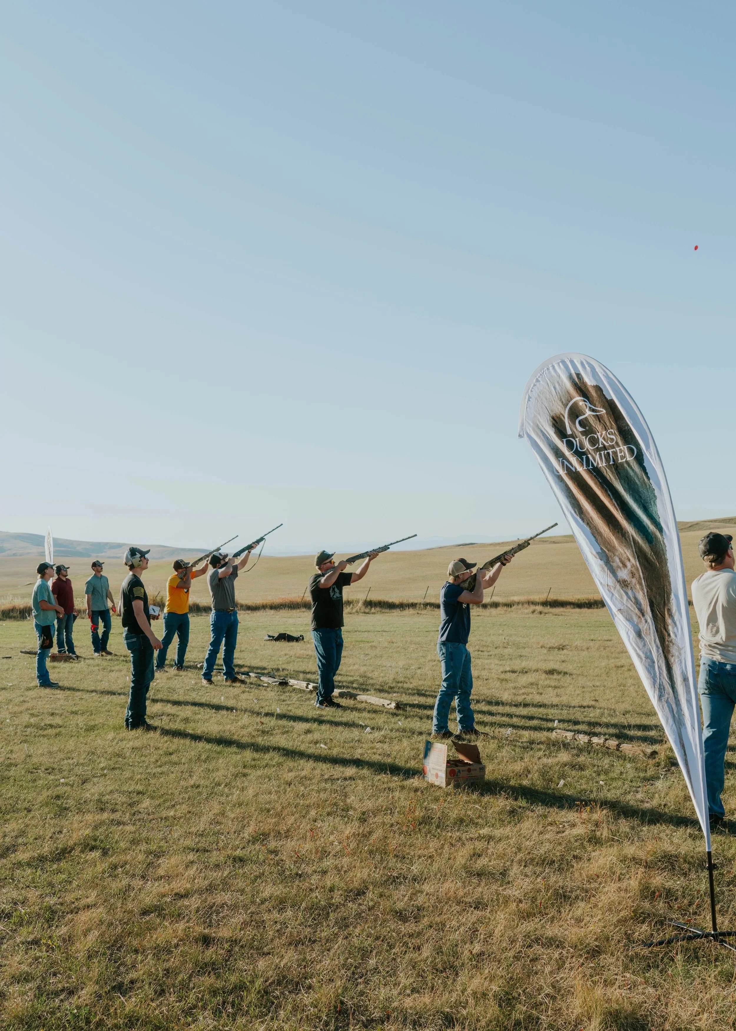 Group of people aiming rifles at a shooting range outdoors, with a large banner saying 'Ducks Unlimited' nearby on a grassy field under clear blue sky.
