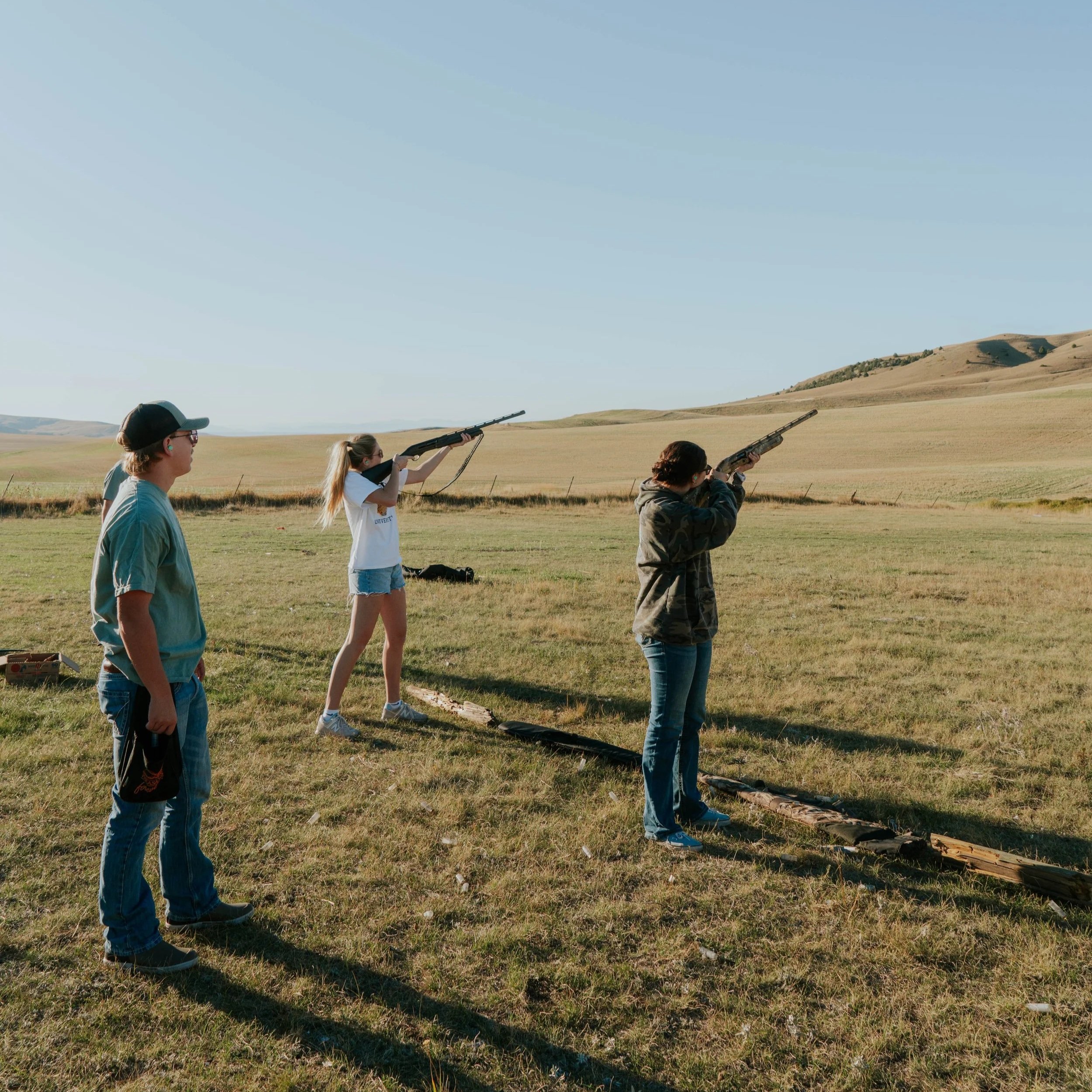 Four people shooting rifles in an open field with rolling hills in the background.