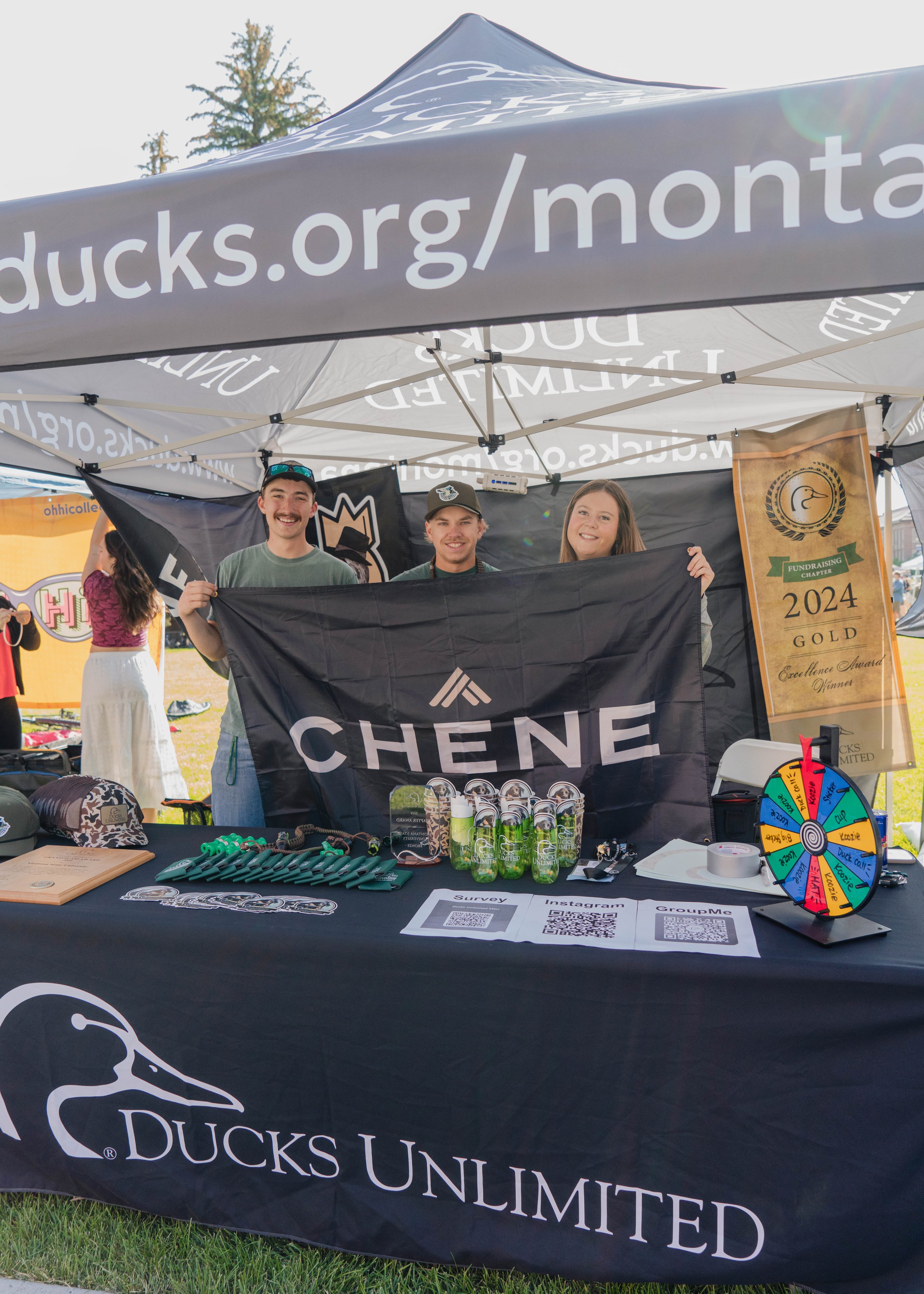 Three people standing behind a table under a canopy tent, holding a black flag with 'CHENE' written on it, at an outdoor event.
