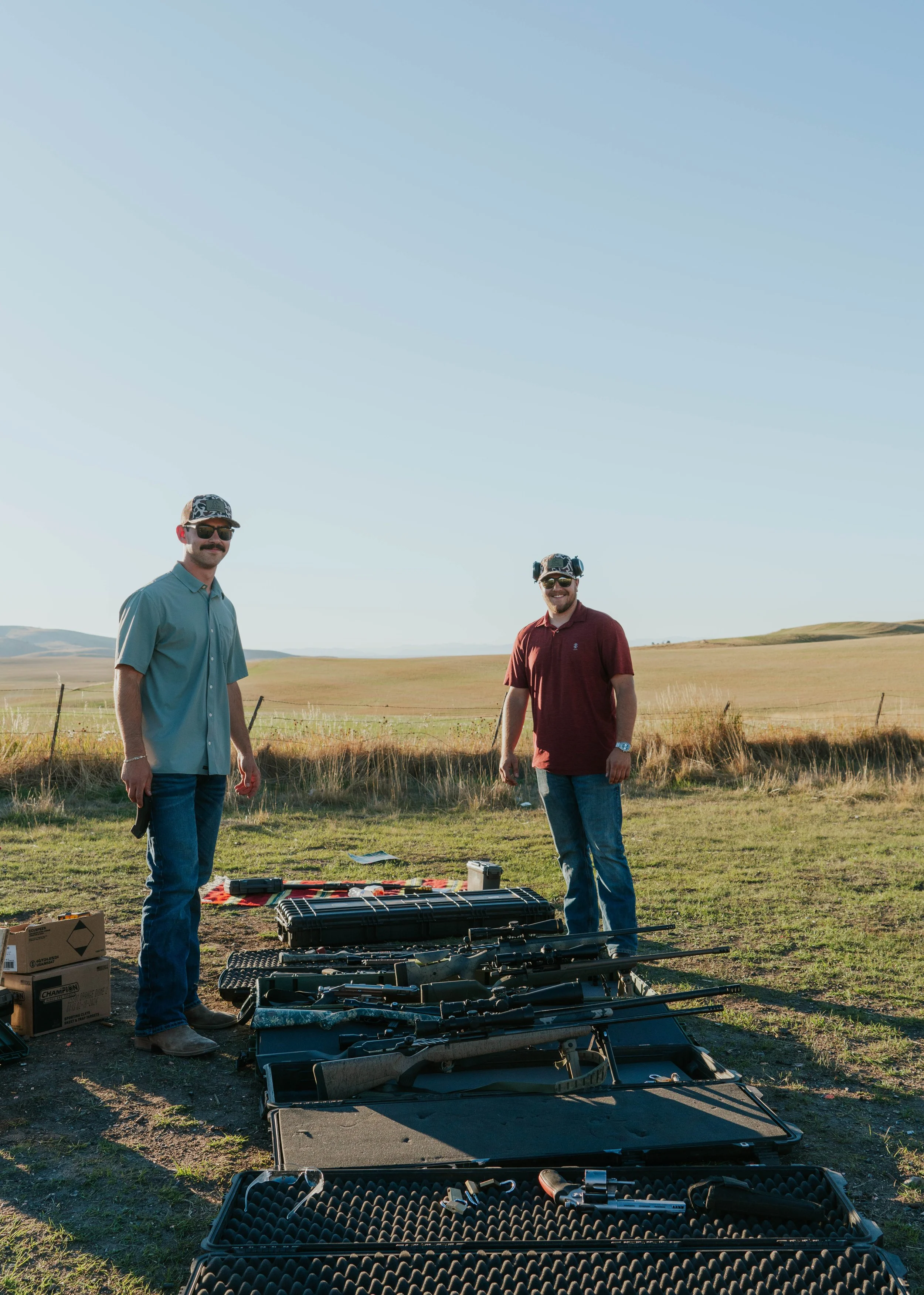 Two men standing outdoors in a field near a table with rifles and equipment, sunny weather, open landscape in the background.