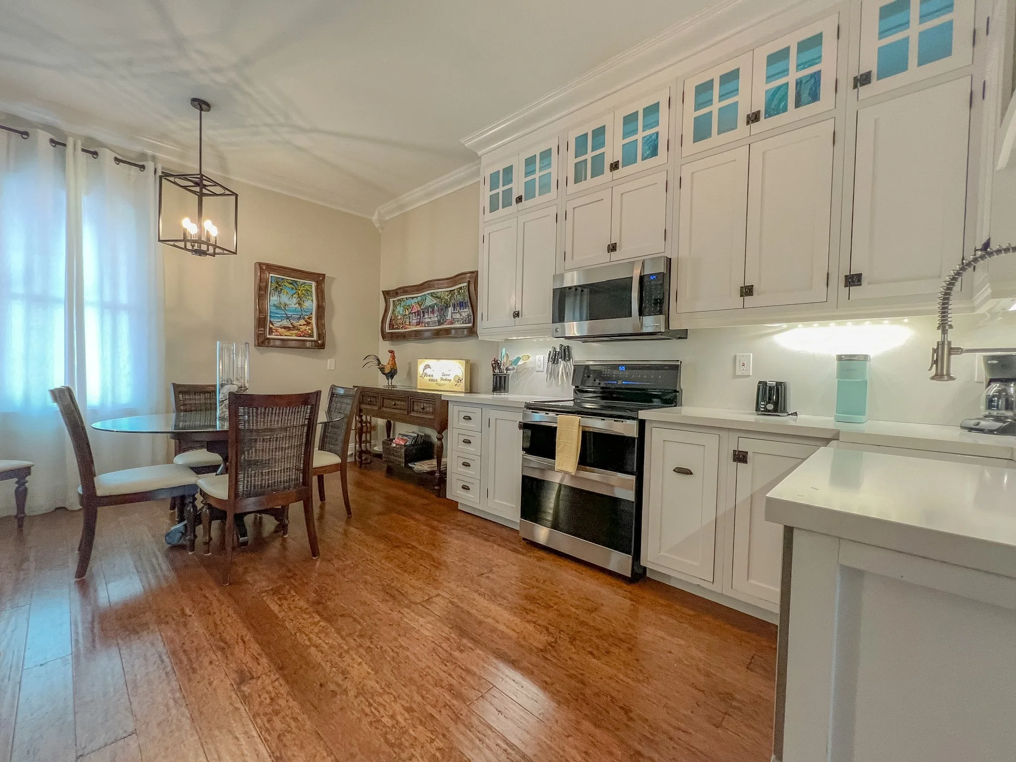 Kitchen with white cabinets, stainless steel appliances, and a wooden dining table with chairs, hardwood floor, and light coming through a large window with white curtains.