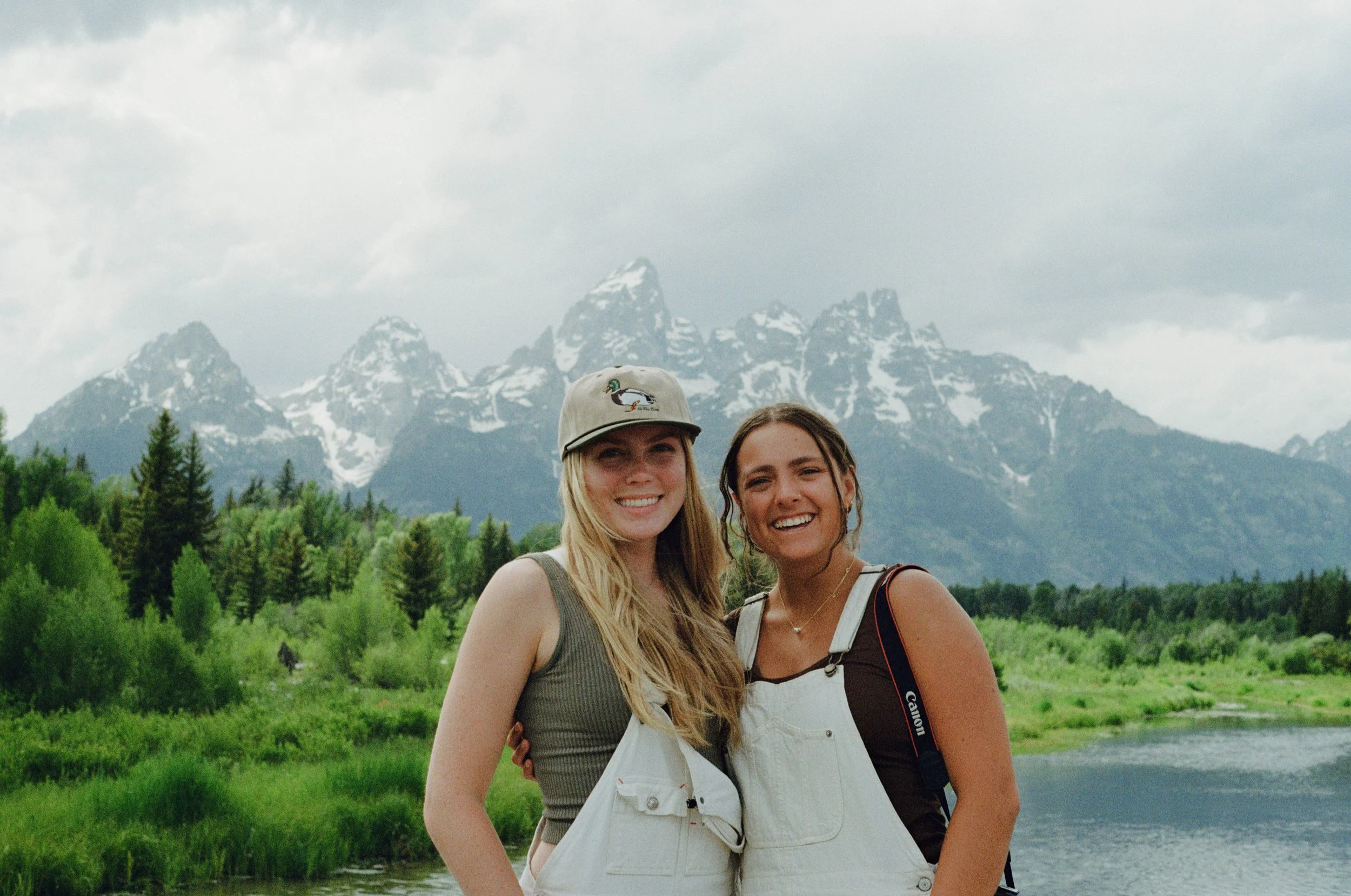 Two young women smiling in front of mountains and a lake, outdoors, on a cloudy day.