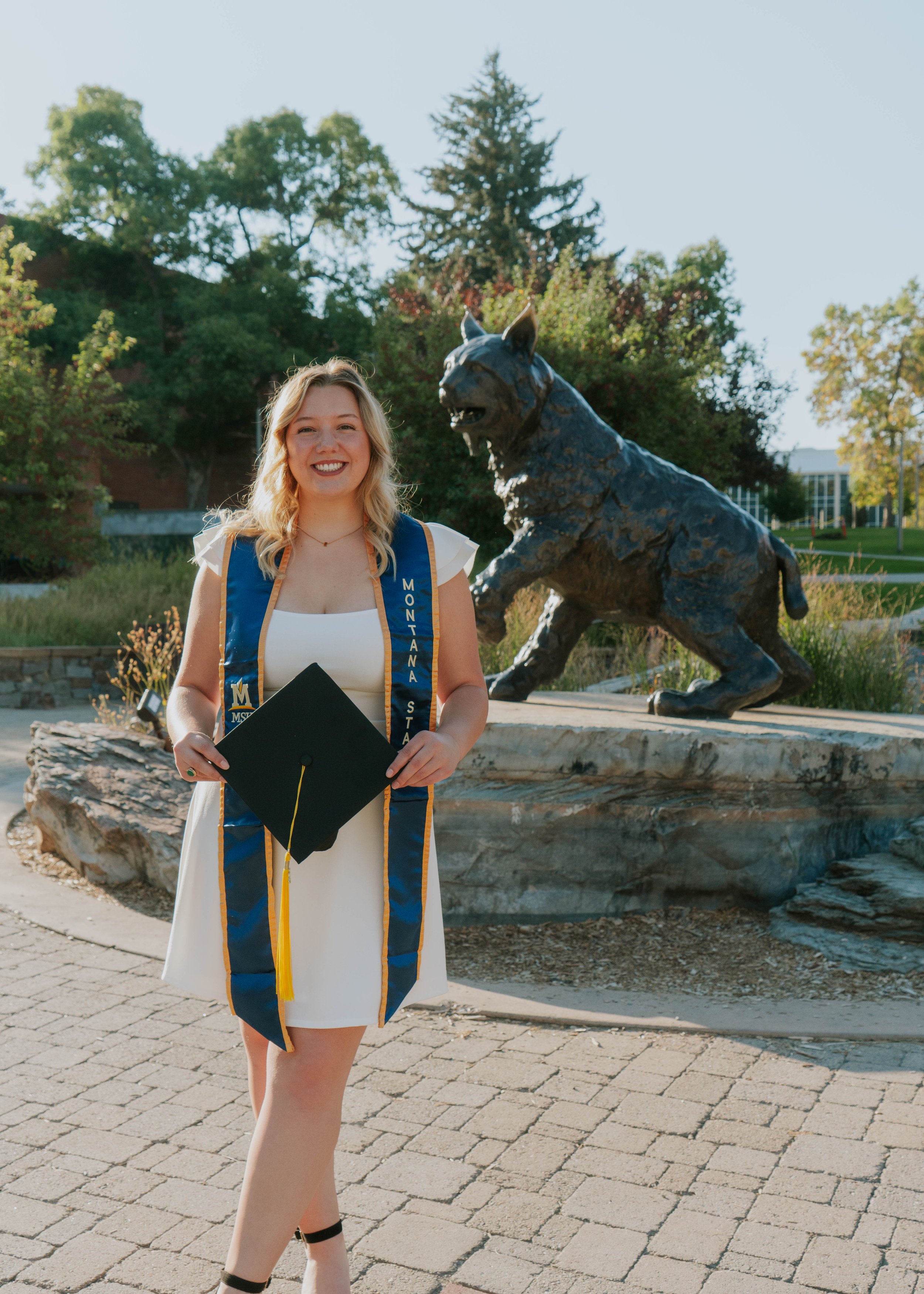 A young woman in a white dress and a blue graduation sash stands outdoors holding a black graduation cap with a yellow tassel. She is smiling and standing in front of a bronze statue of a fierce-looking wolf on a stone base, with green trees and a building in the background.