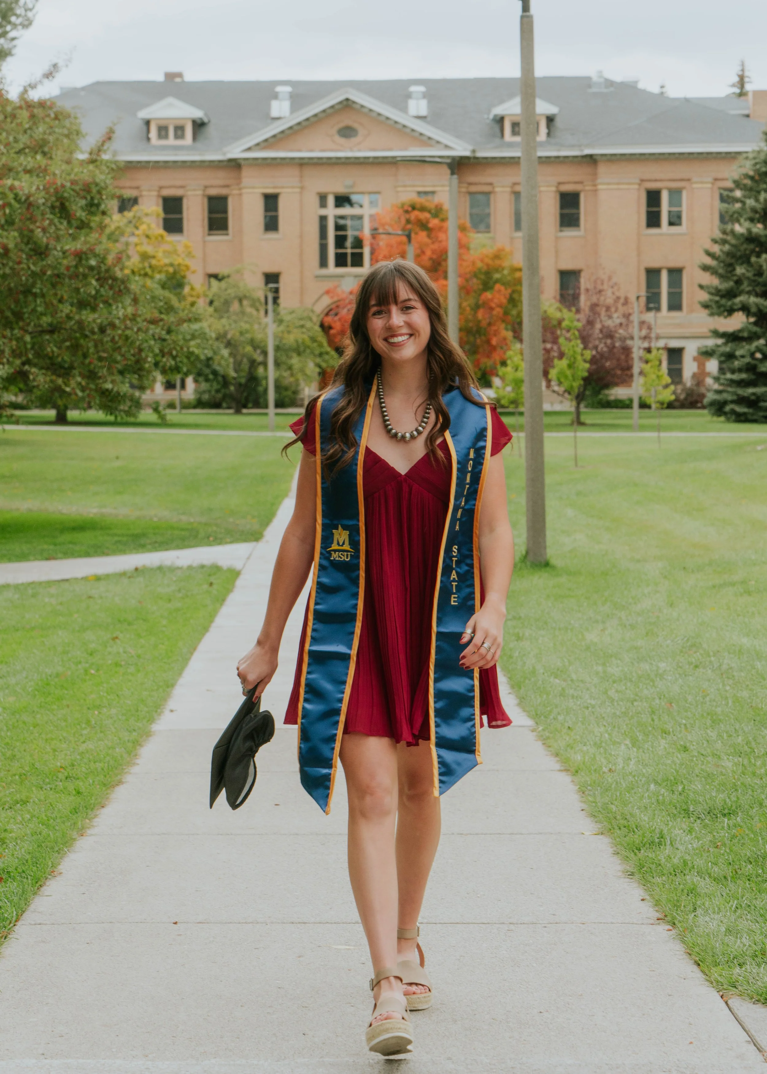 A young woman in a red dress and graduation gown walking on a campus sidewalk, holding a graduation cap in her hand, smiling after graduation.