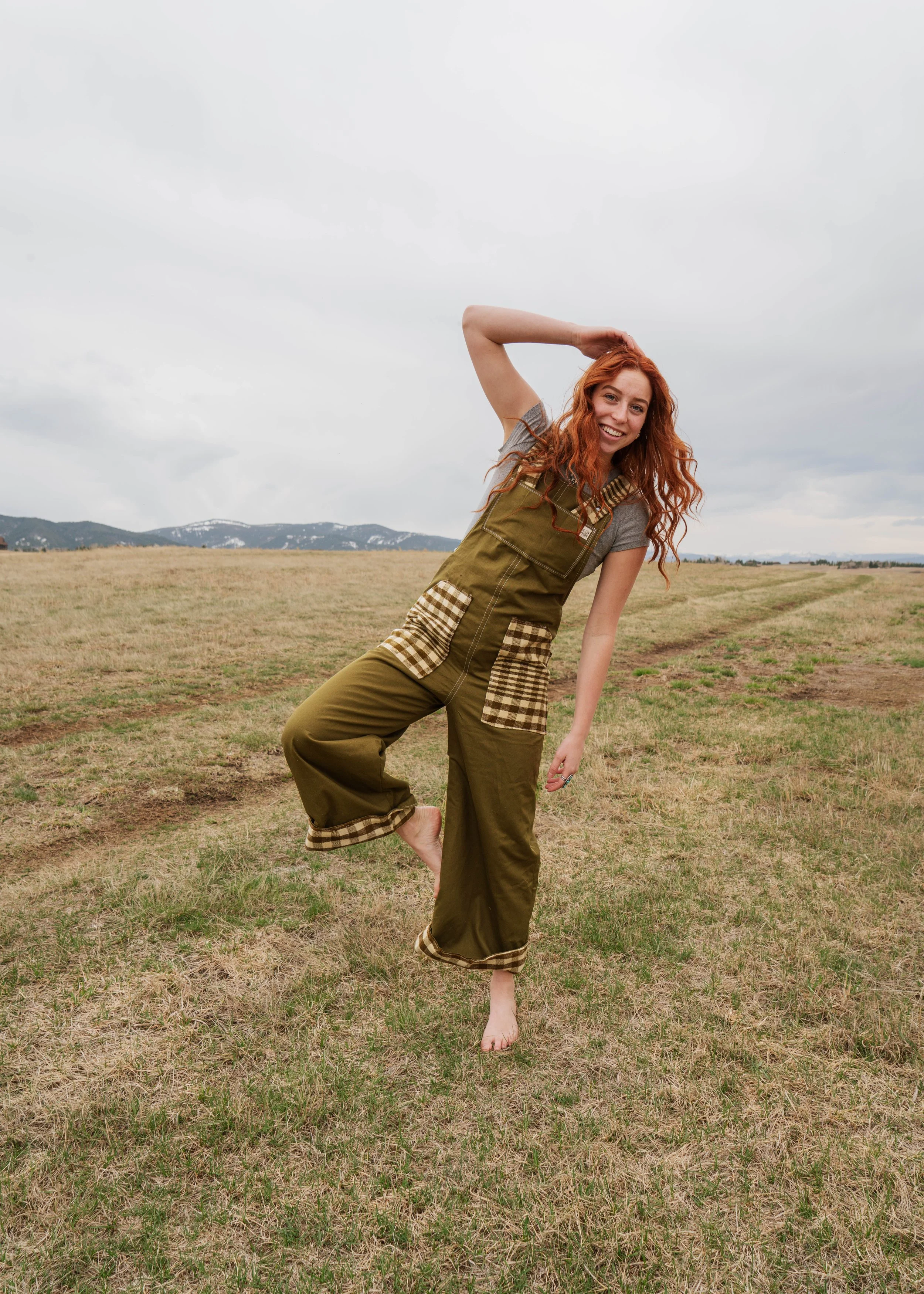 A young woman with curly red hair smiling and balancing on one foot in a grassy open field, dressed in green overalls with plaid pockets and trim, with mountains and cloudy sky in the background.