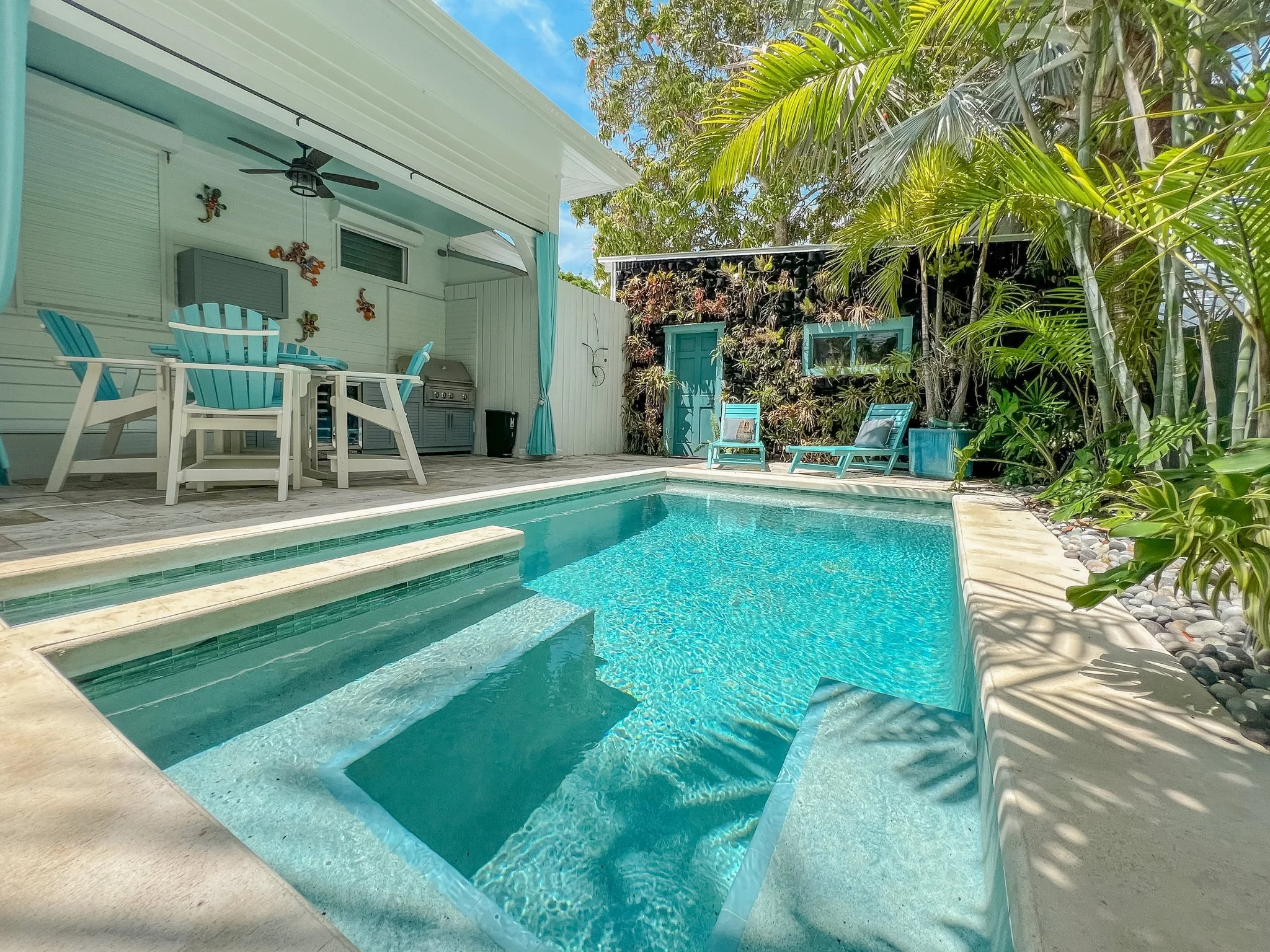 A backyard swimming pool surrounded by tropical plants, with a covered patio area on one side featuring a dining table, chairs, and a barbecue grill. Two lounge chairs are on the far side of the pool, and the sky is clear with some clouds.