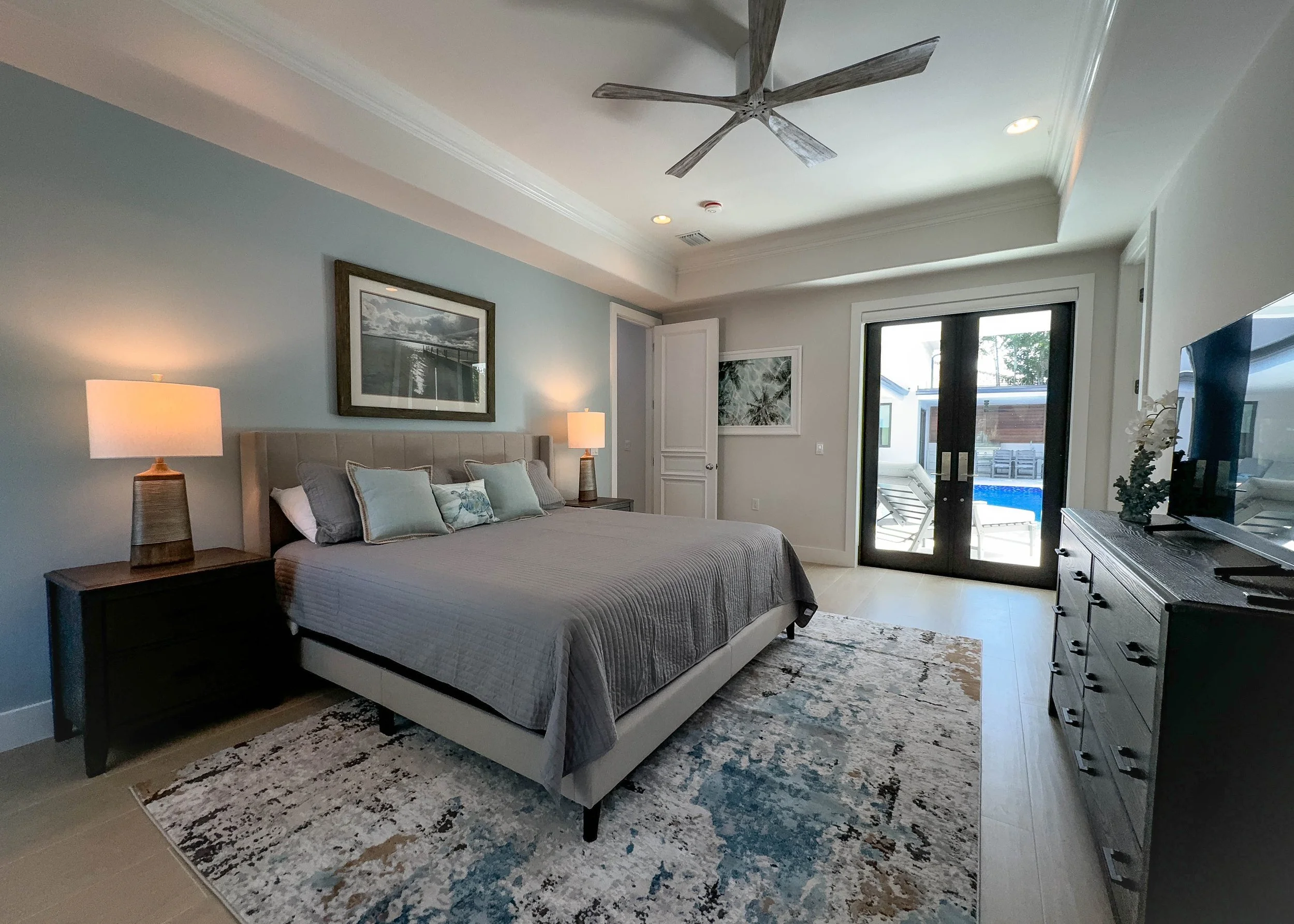 A modern bedroom with a large bed, nightstands with lamps, a dresser with a TV, and a sliding glass door leading to an outdoor pool area.