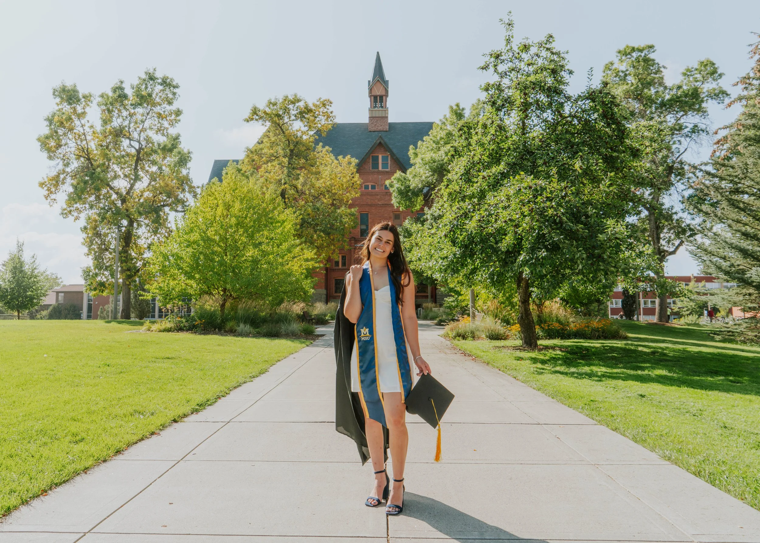 A young woman in a white dress and black heels standing on a college campus sidewalk, holding a diploma and graduation cap, wearing a graduation stole, with a red brick university building and trees in the background on a sunny day.