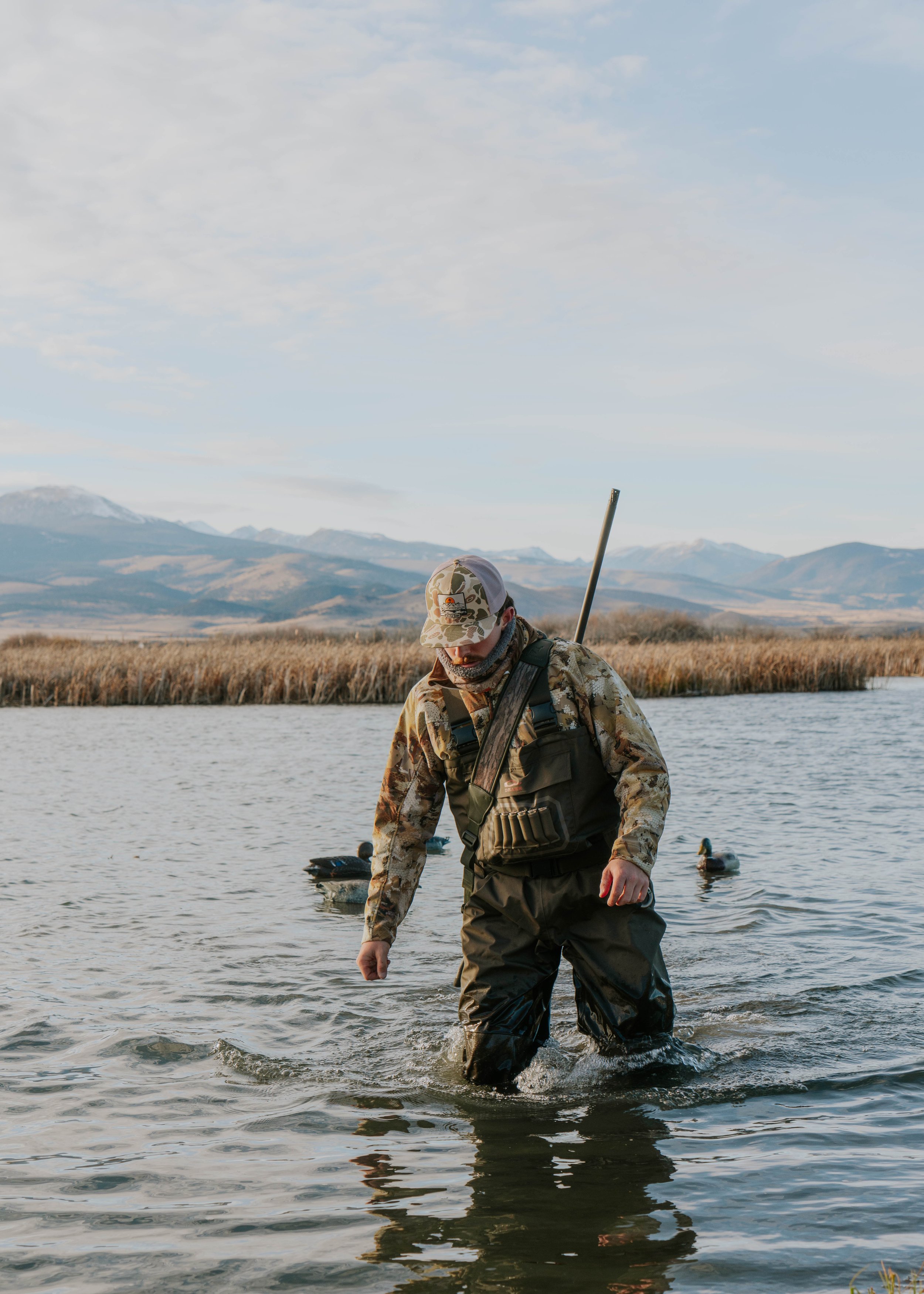 A person in camouflage clothing and waterproof gear wading through a lake with ducks in the background and mountains in the distance.