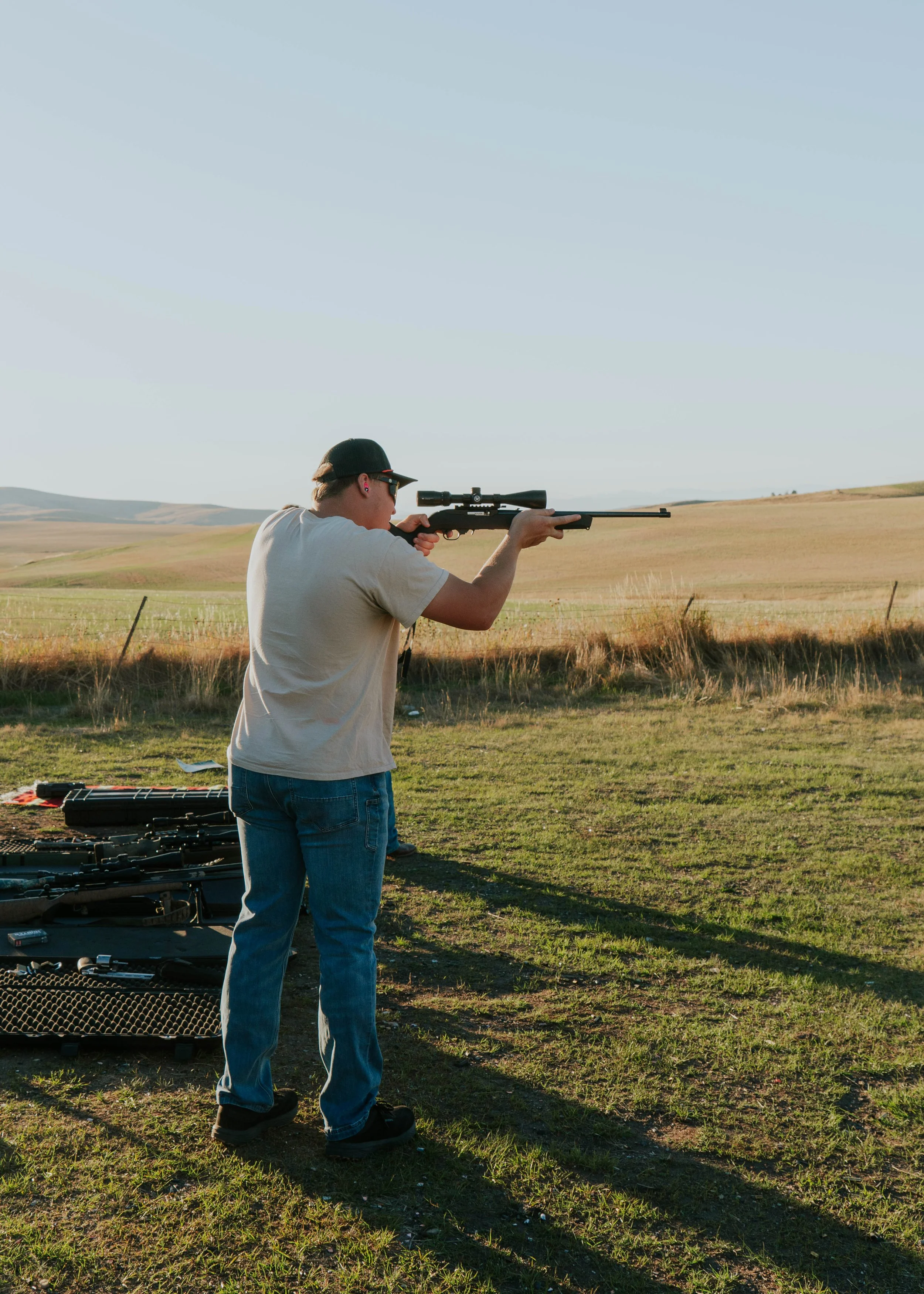 A man wearing a black cap, sunglasses, a beige t-shirt, and jeans is aiming a rifle with a scope at an outdoor shooting range during the day. There are multiple rifles and accessories on the ground and in cases nearby.