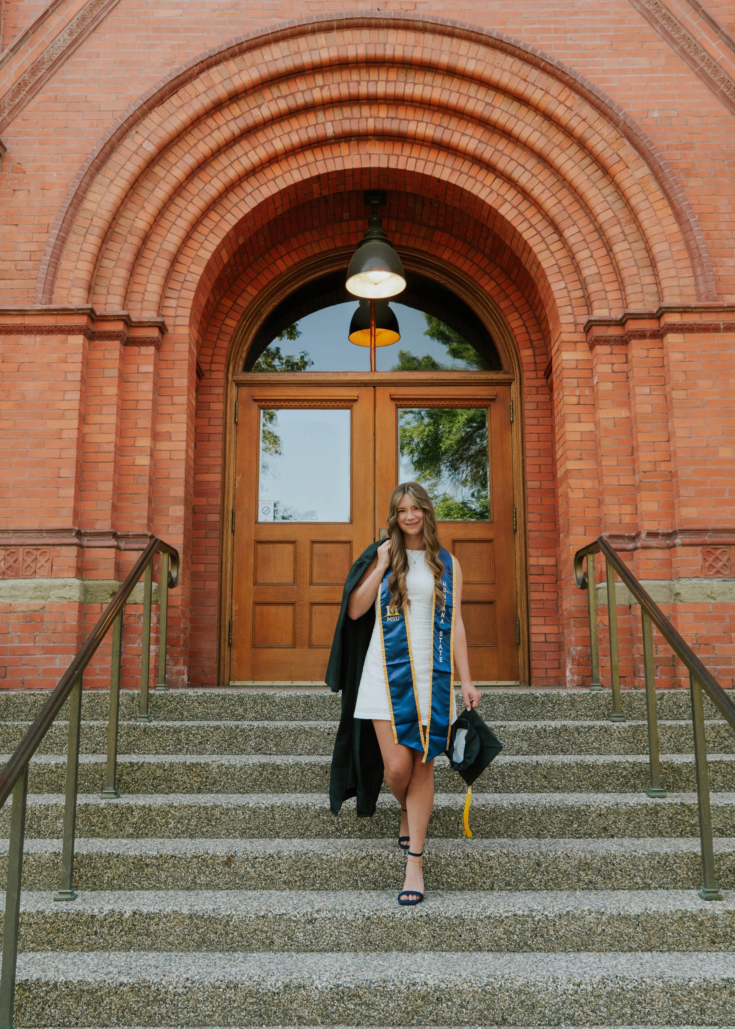 A young woman in a white dress, wearing a graduation stole and cap, stands on the steps of a brick building after graduation, smiling and carrying her gown and cap.