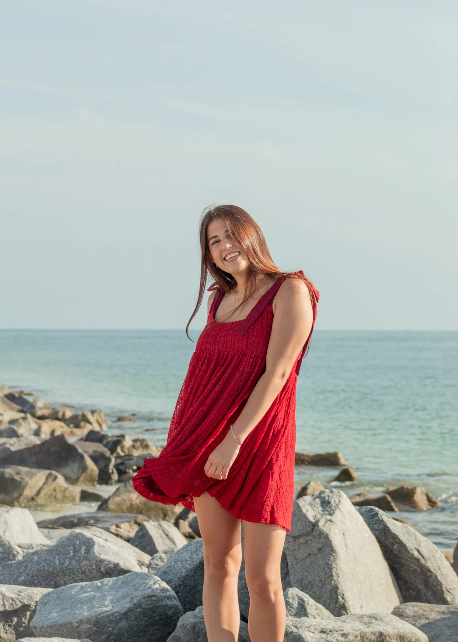 A young woman in a red dress standing on rocks by the ocean, smiling and looking at the camera.