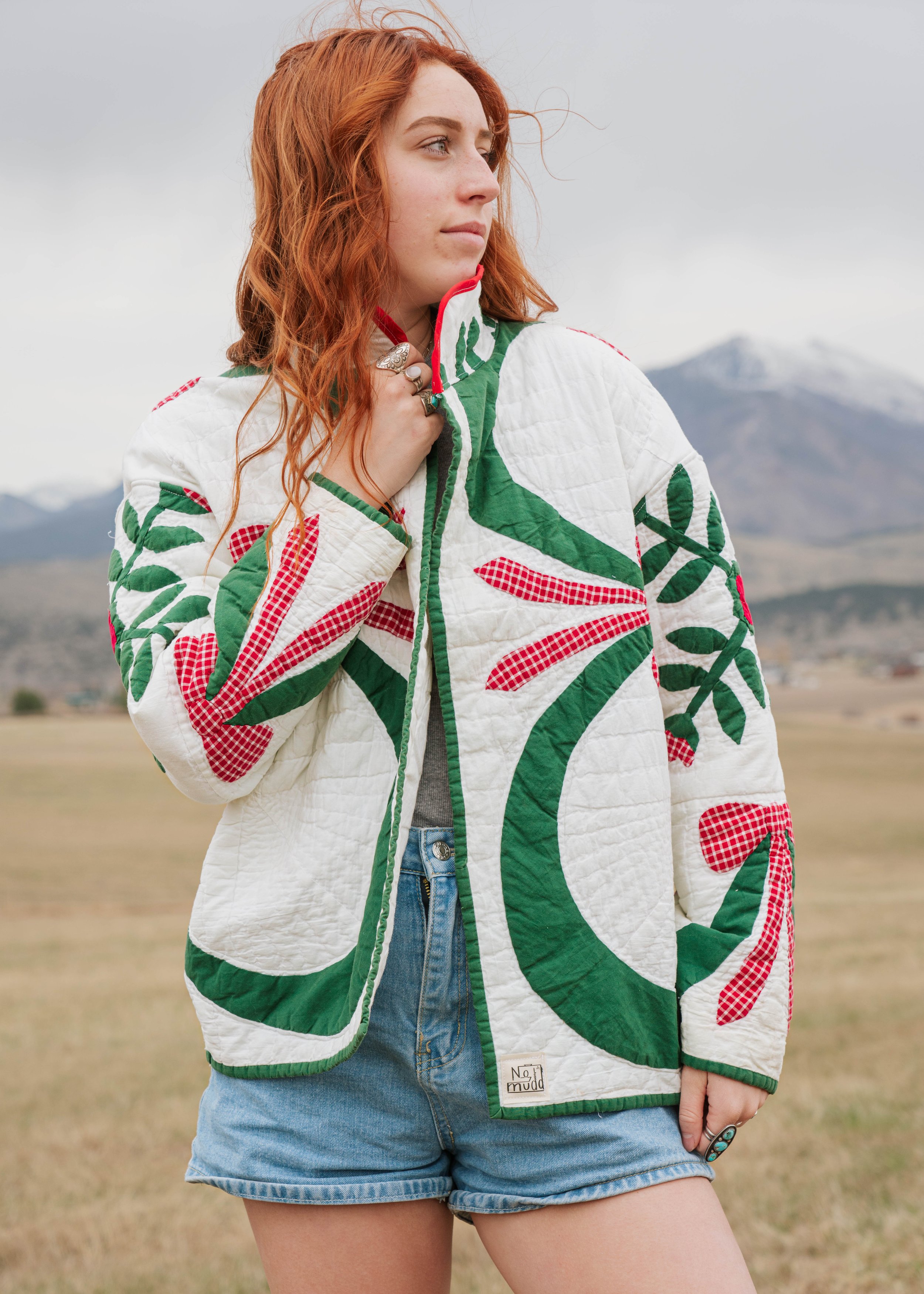 A woman with long red hair wearing a colorful quilted jacket with green, red, and white patterns, standing outdoors with mountains in the background.