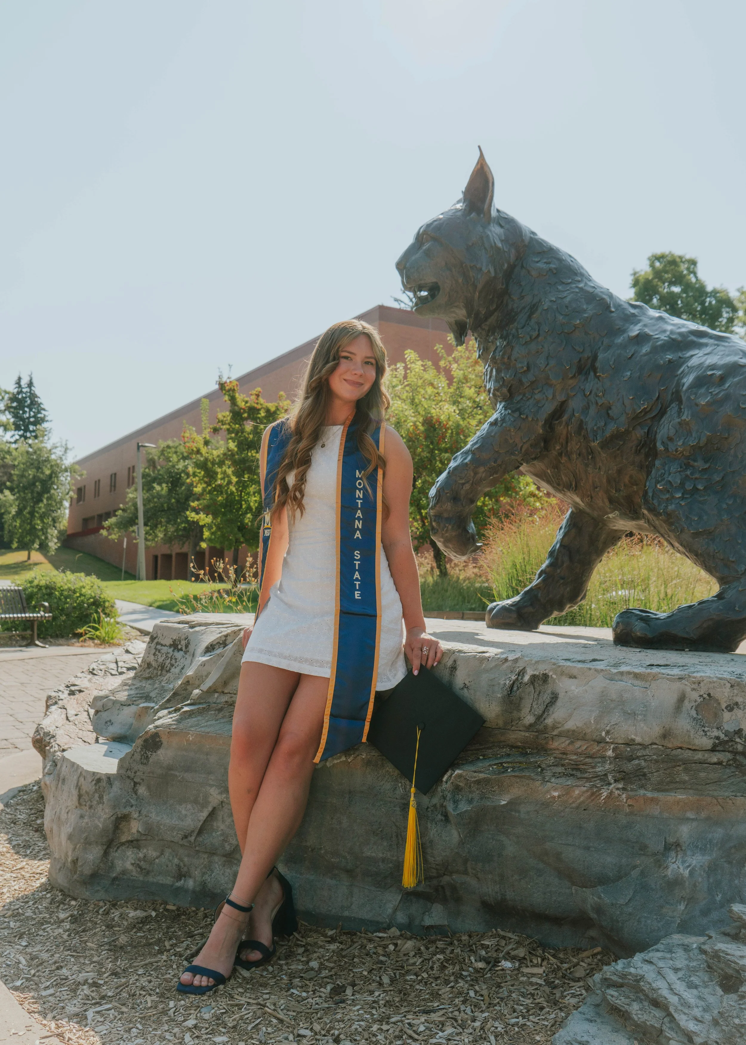 Young woman in white dress and black heels holding a graduation cap, standing next to a bronze mountain lion statue outdoors.