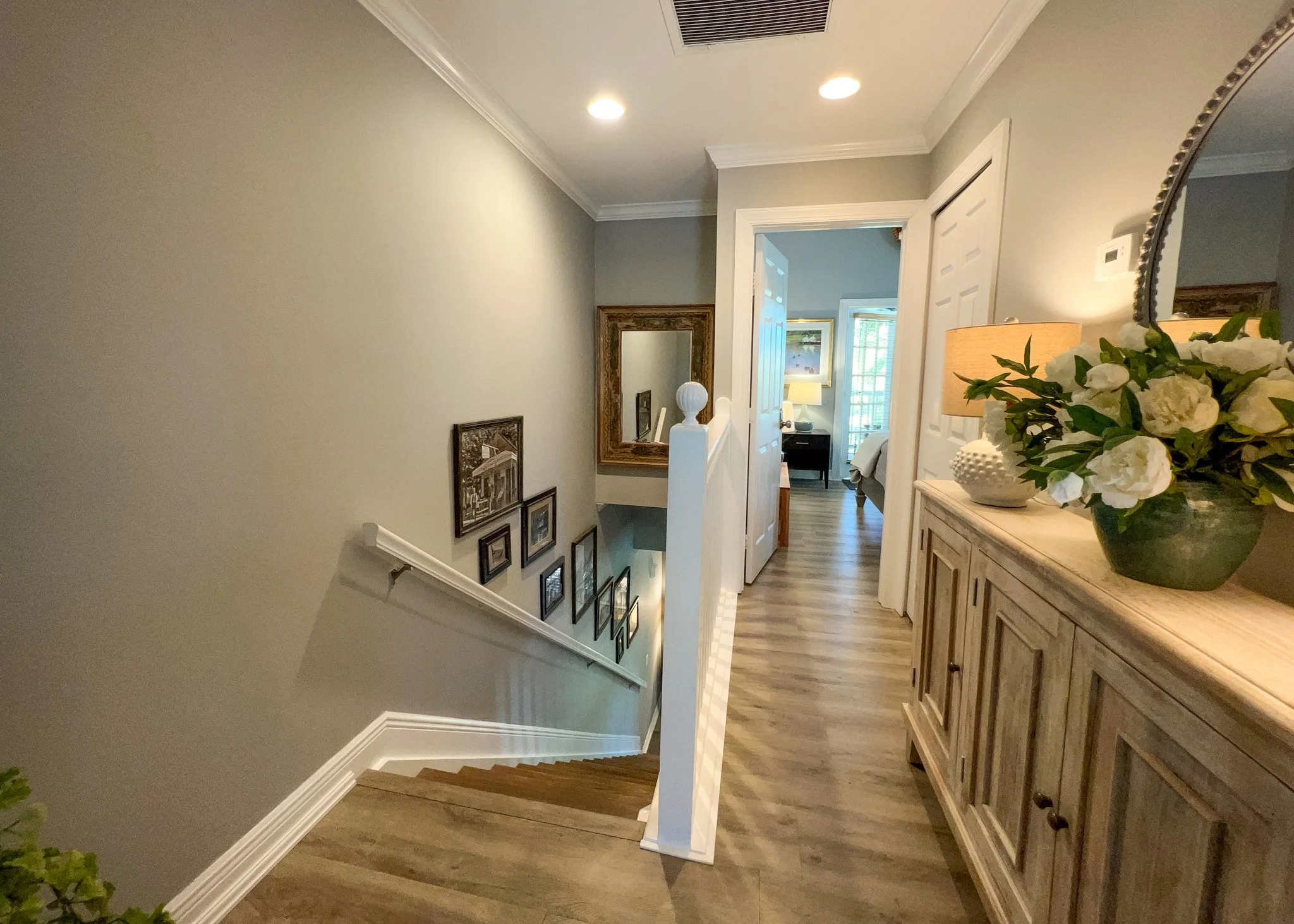 Interior view of a home hallway leading to a bedroom, featuring a staircase with framed pictures on the wall, a wooden sideboard with a lamp, and a large flower arrangement, with natural light coming through the windows.