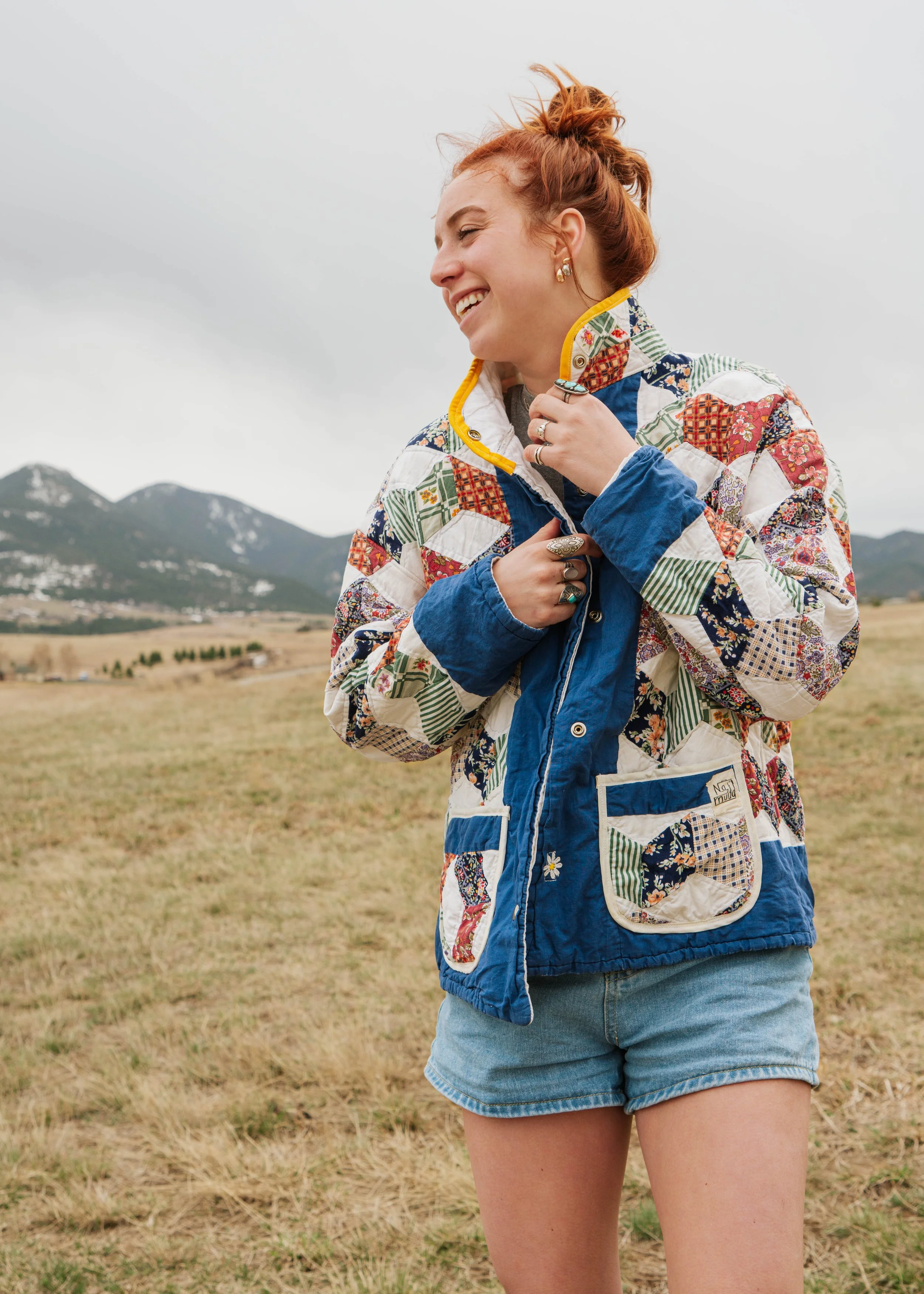 A woman with tousled red hair smiling outdoors, wearing a colorful quilted jacket with patchwork design and blue denim shorts, standing in a grassy field with mountains in the background.