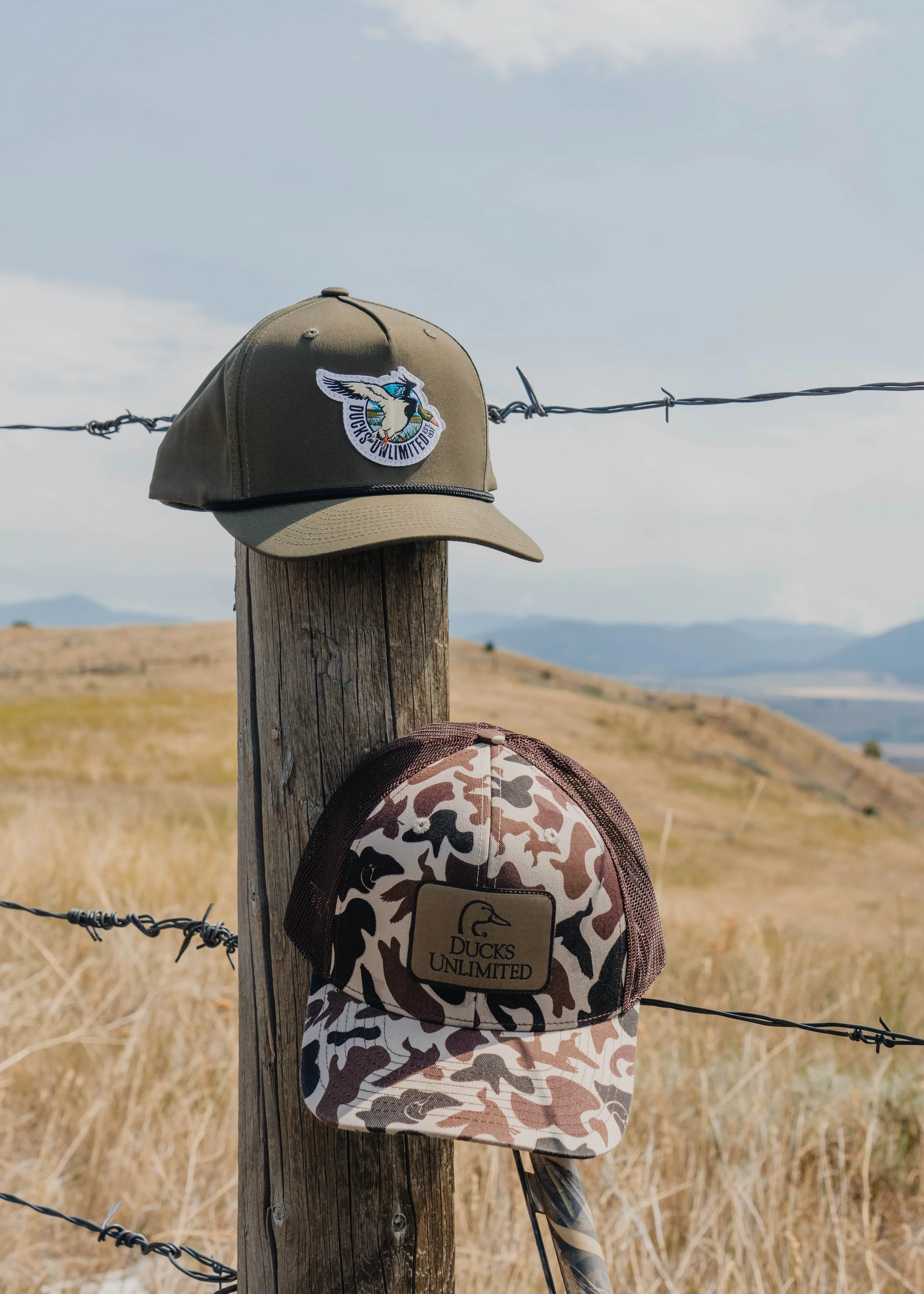 Two hats hanging on a wooden fence post in a rural landscape, one green with a bird and the text 'Ducks Unlimited,' the other camouflage with an embroidered patch and the text 'Ducks Unlimited.'