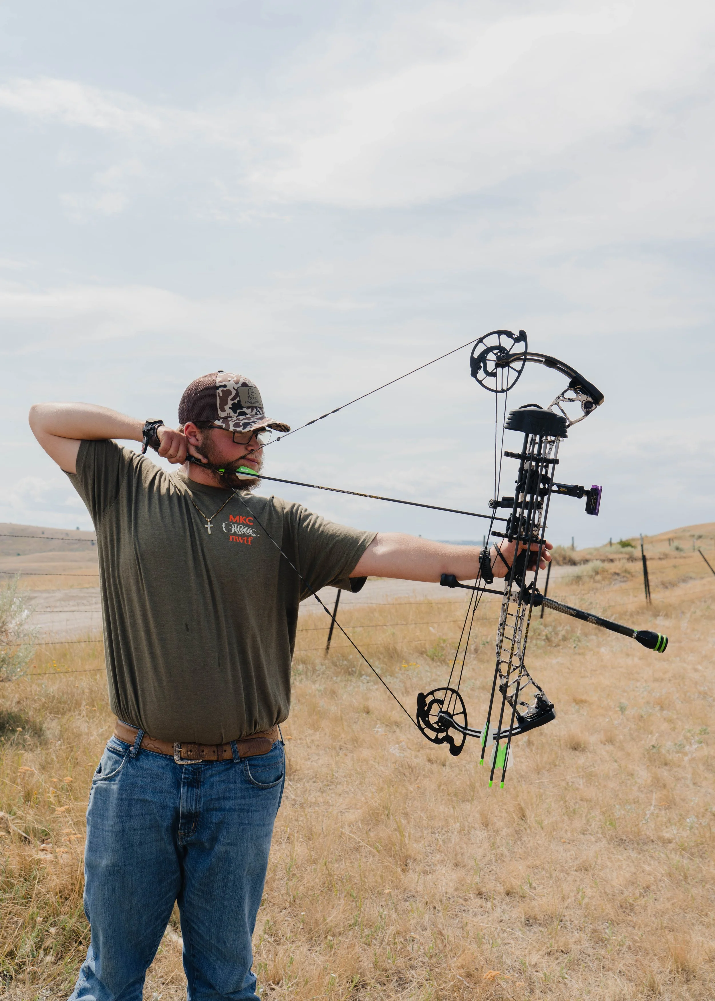 A man standing outdoors in a field aiming a compound bow, with the bow drawn back and ready to shoot.