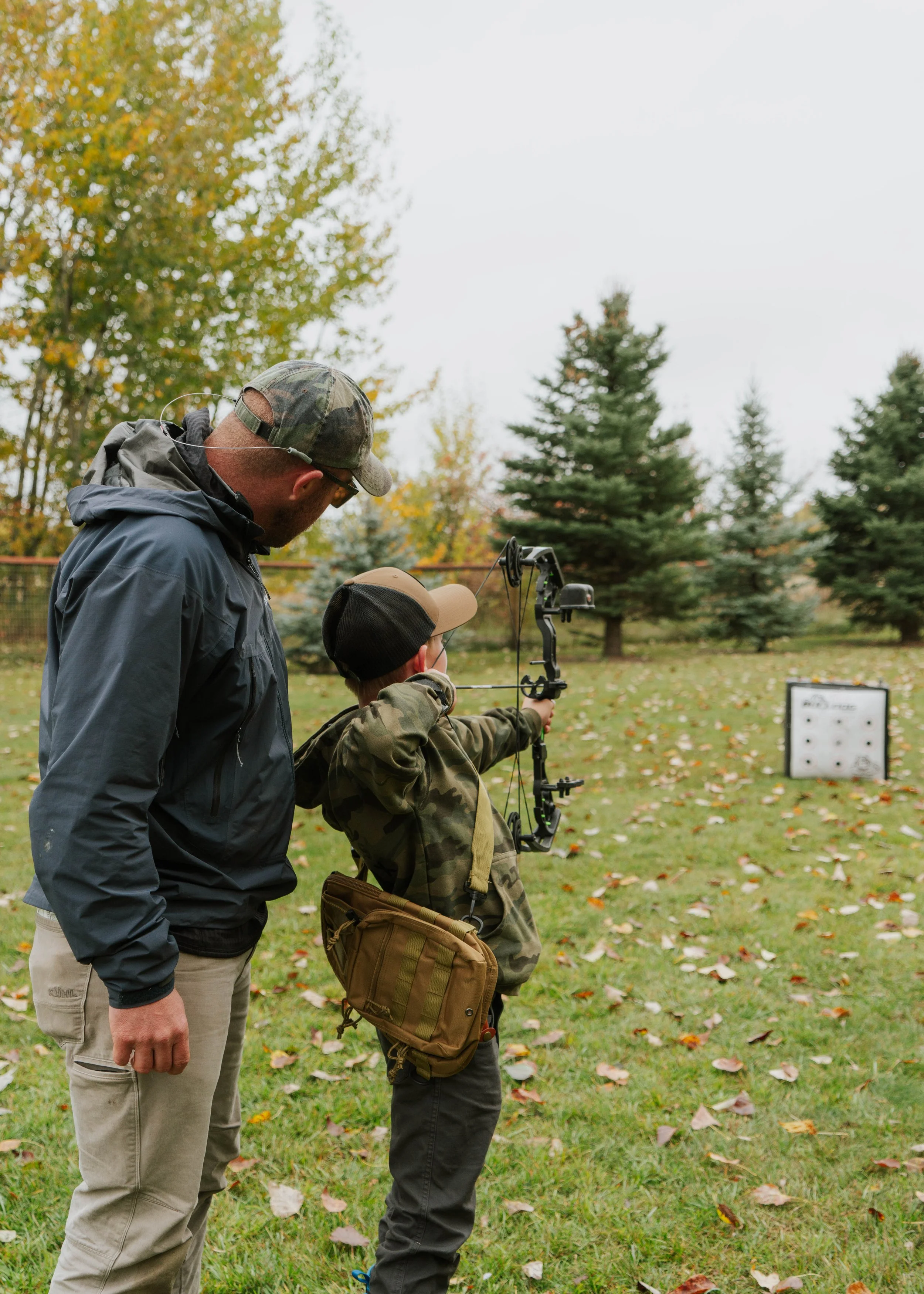 A man is coaching a young boy how to shoot a bow and arrow outdoors on a grassy field with trees and fall leaves.
