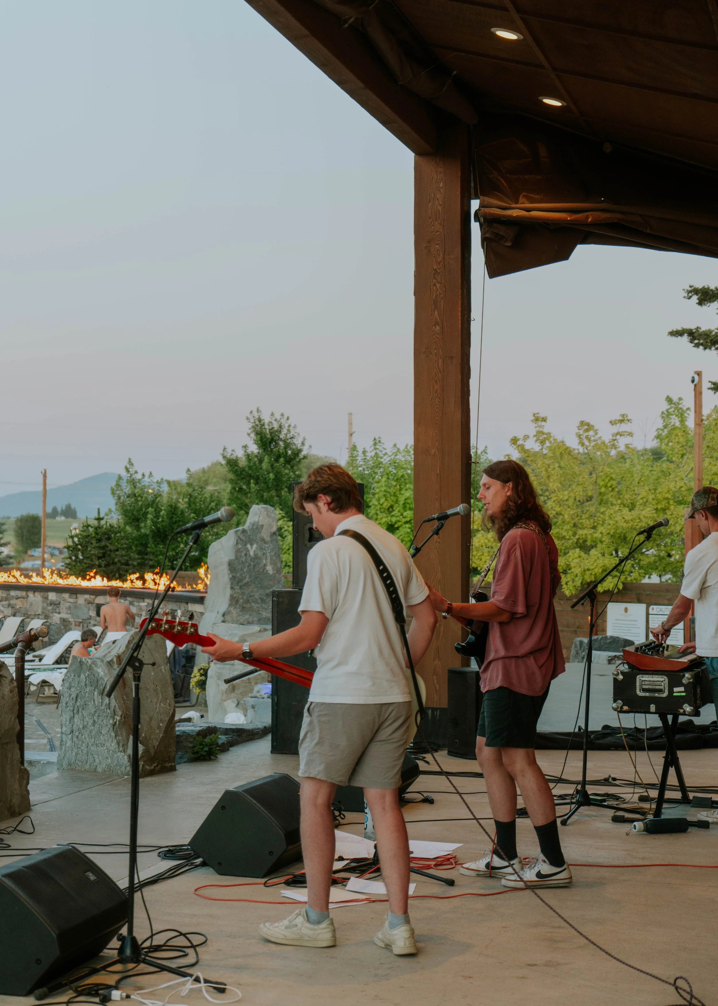 A band performing on an outdoor stage overlooking a pool and a scenic landscape with trees and mountains. The band consists of two guitarists, one in a white shirt and shorts and the other in a brown shirt and shorts, with a DJ or sound technician in the background. The setting appears to be at sunset or dusk.