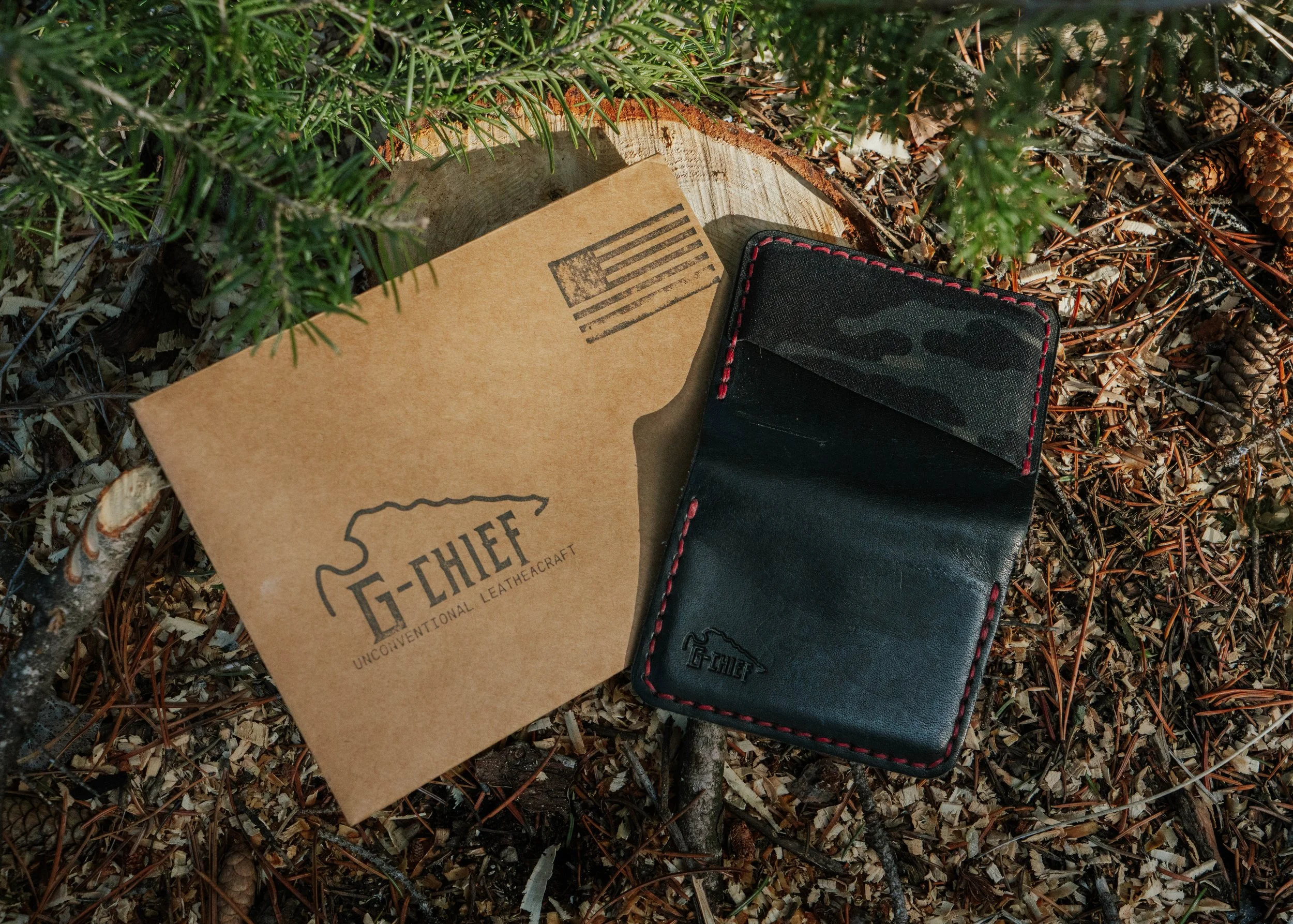 A black leather wallet with red stitching resting on forest ground next to a cardboard piece with a G-THIEF logo and slogan, surrounded by pine needles, small twigs, and pine cones.