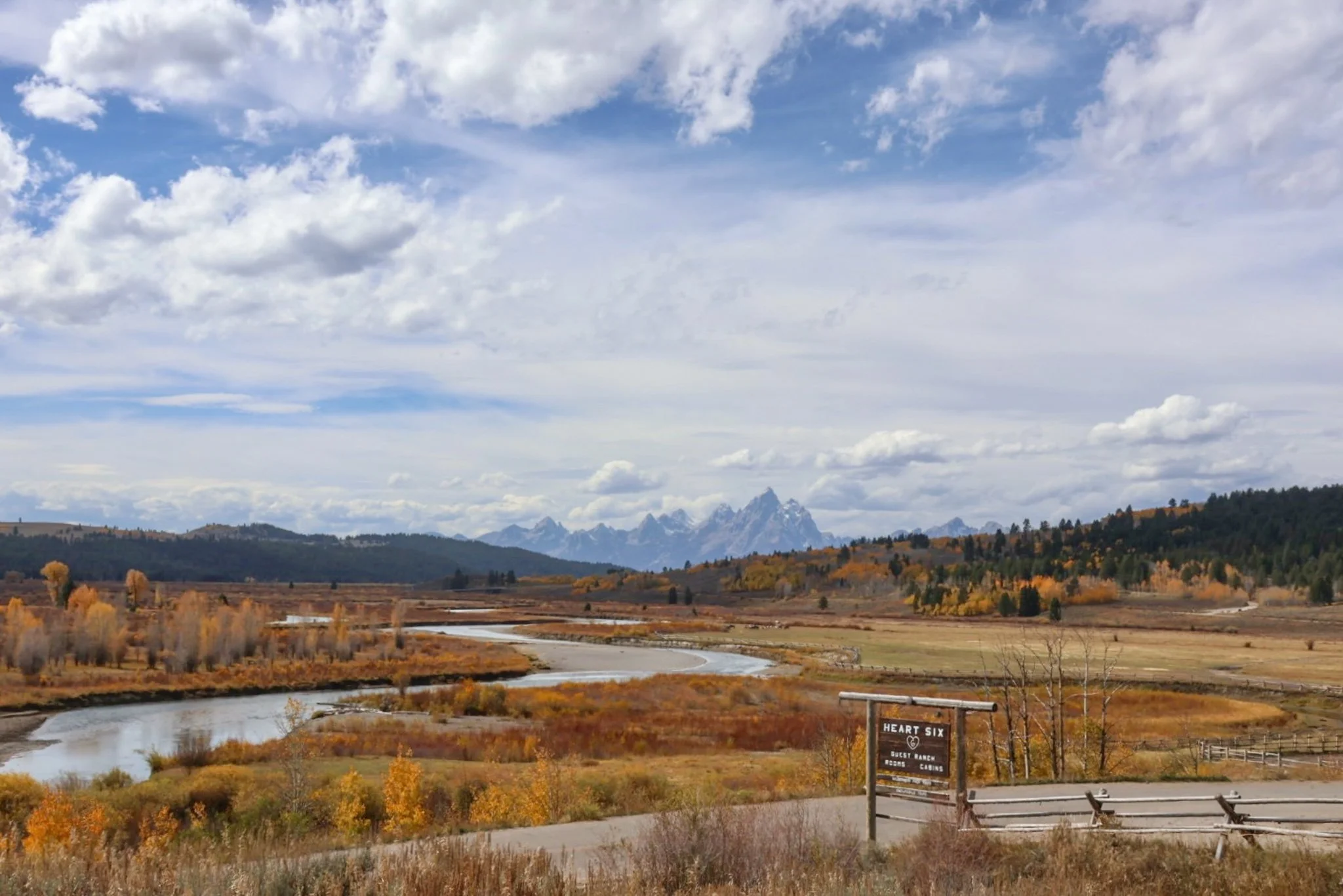 A scenic view of a river meandering through a landscape with fall foliage, mountains in the distance, and a cloudy sky overhead. There is a sign in the foreground that says 'Heart Six Guest Ranch, Rooms, Cabs'.