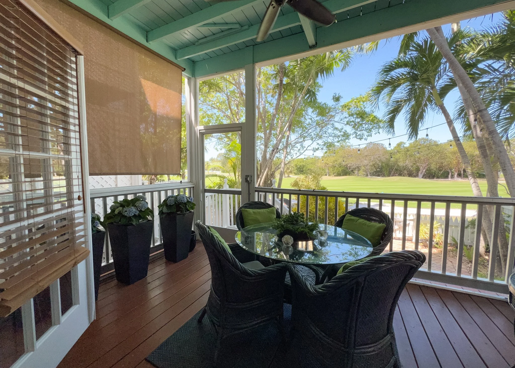 A screened porch with a glass-top table and wicker chairs, overlooking a lush green golf course with palm trees, under blue sky with string lights hanging above.