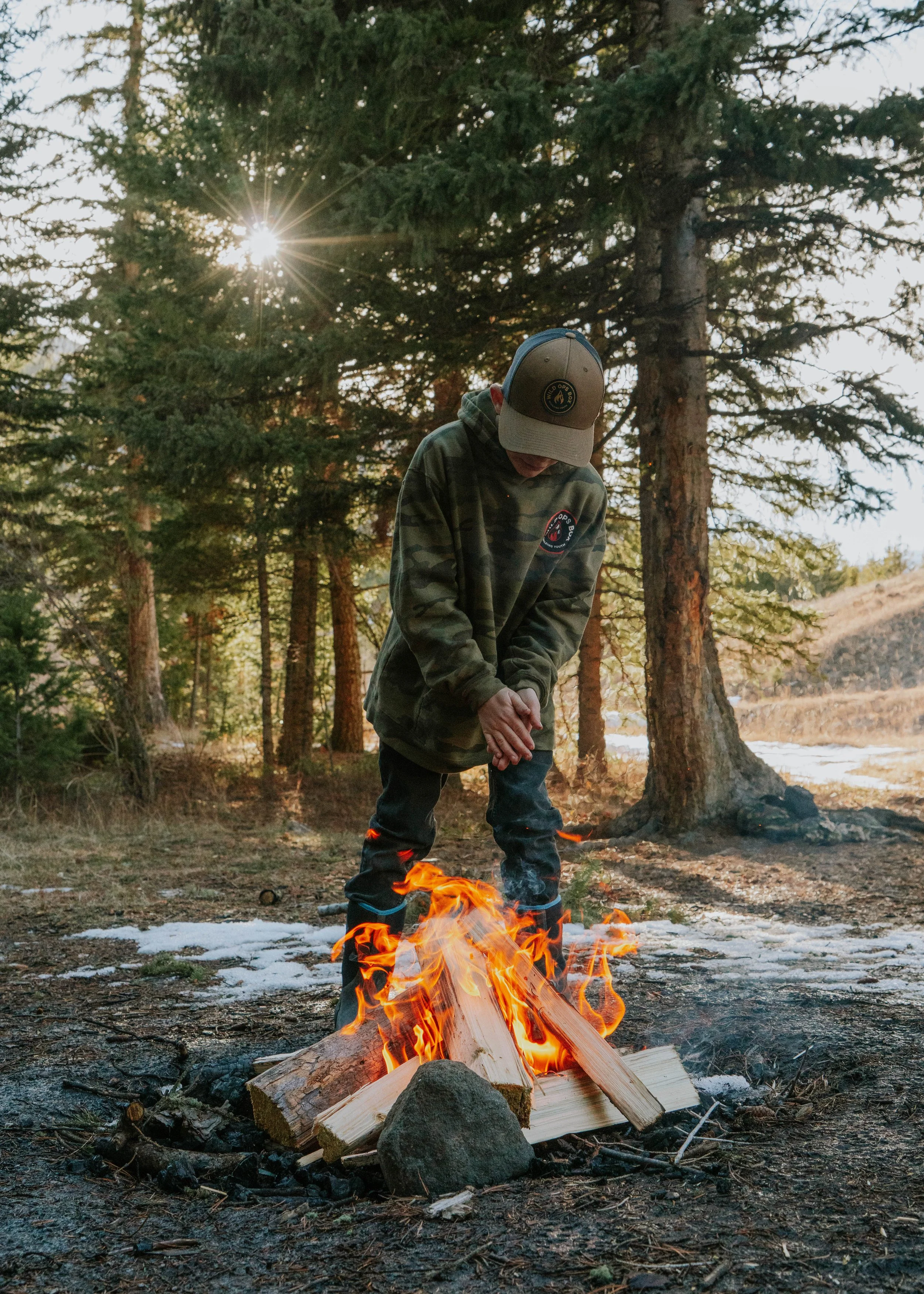 A person in camouflage hoodie and cap standing near a campfire in a forested area during daytime.