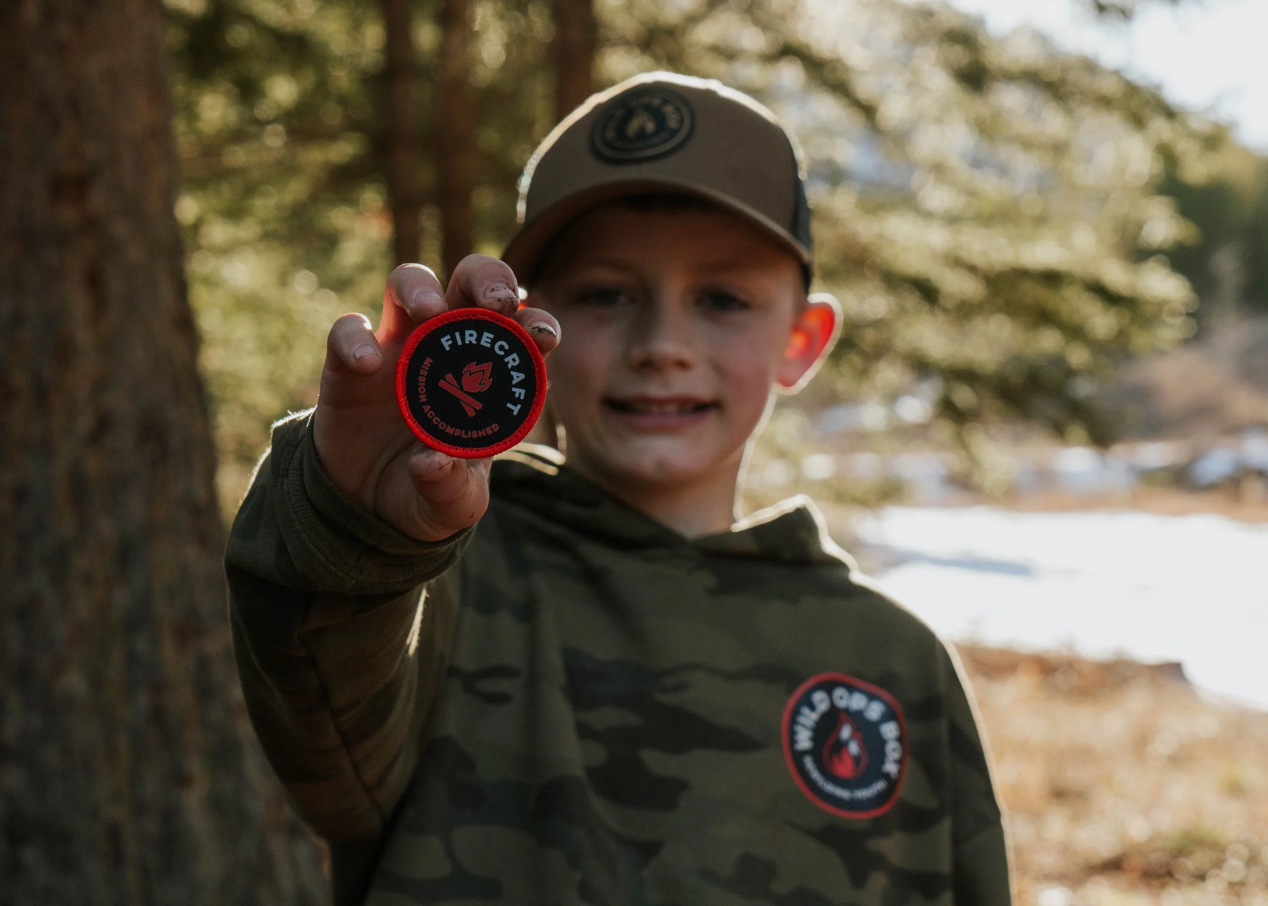A young boy outdoors in a wooded area, wearing a camouflage hoodie with a Boy Scout Firecraft badge, holding a Firecraft embroidered patch close to the camera, slightly out of focus.