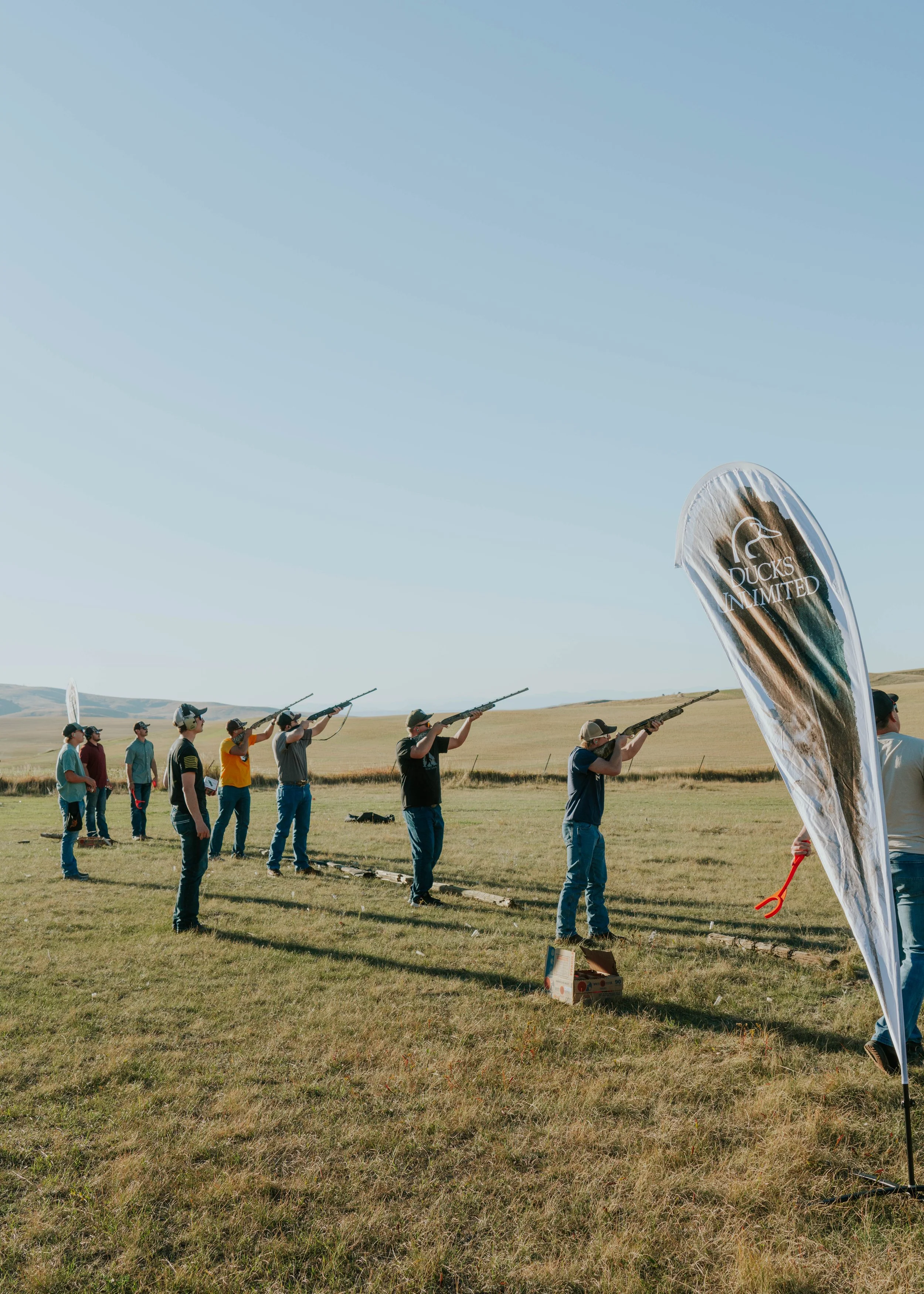 Multiple people participating in a clay pigeon shooting activity outdoors in a grassy field with mountains in the background, some aiming rifles, and a flag with the logo 'Ducks Unlimited'.