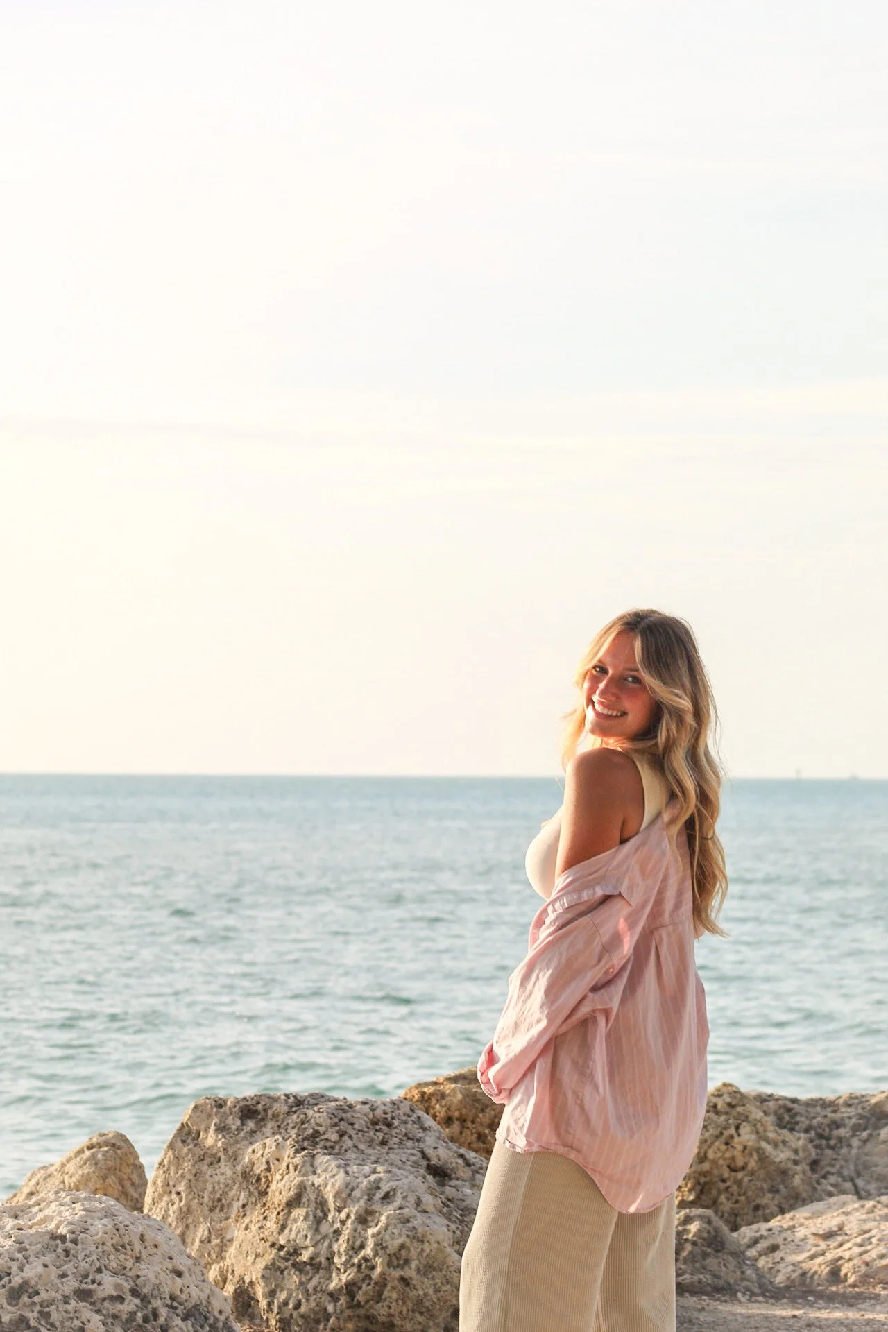 Young woman with long blonde hair smiling at the camera, standing on rocks near the ocean with a clear sky