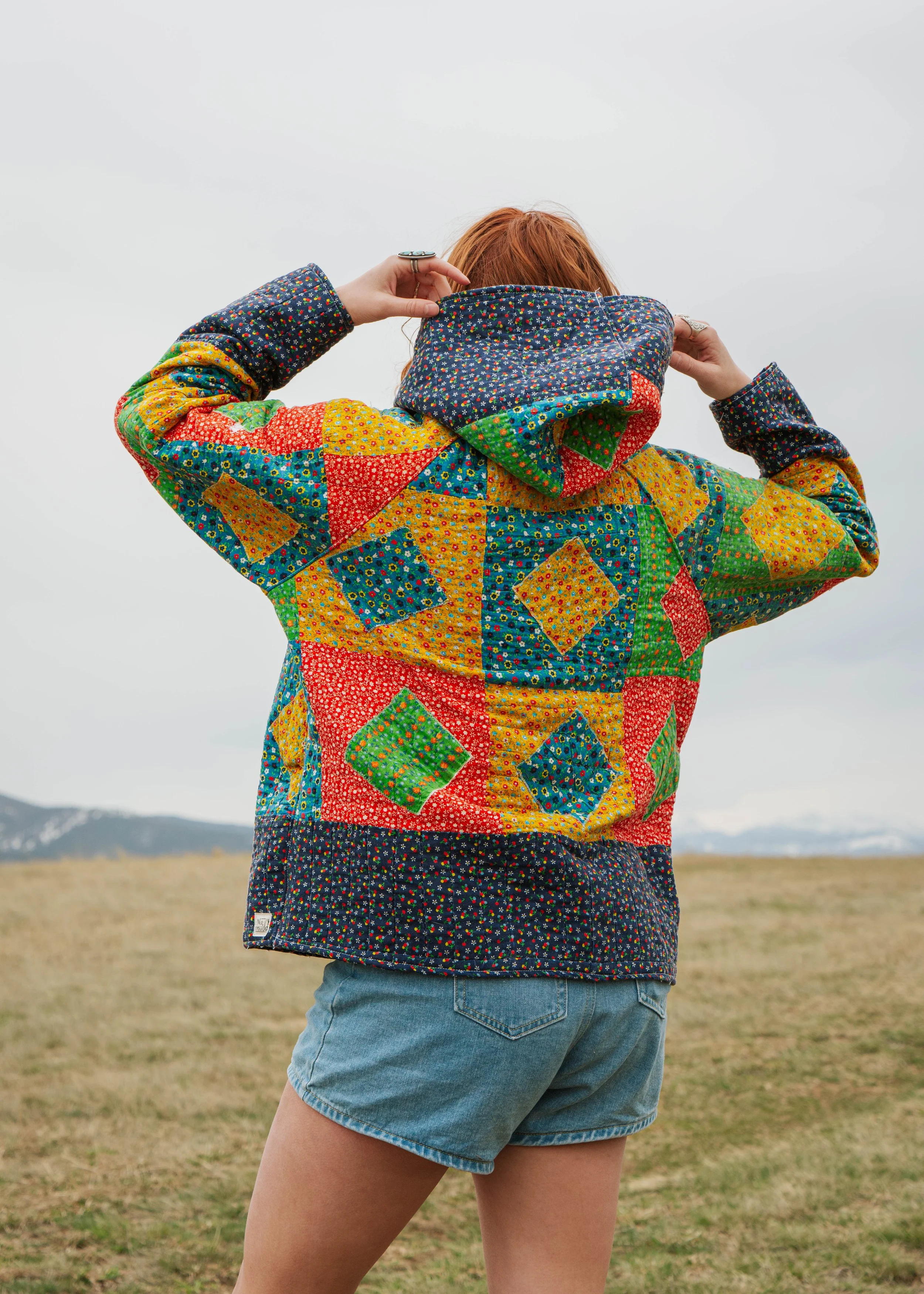Person with red hair wearing a colorful patchwork quilted jacket and denim shorts, standing outdoors in a field with mountains in the background, facing away from the camera and adjusting the hood.