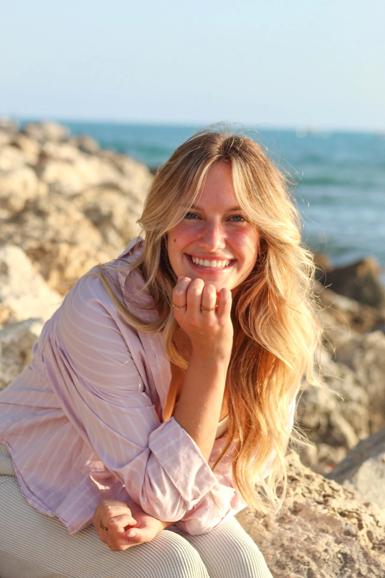 A smiling woman with long blonde hair sitting on rocks near the ocean during the day.