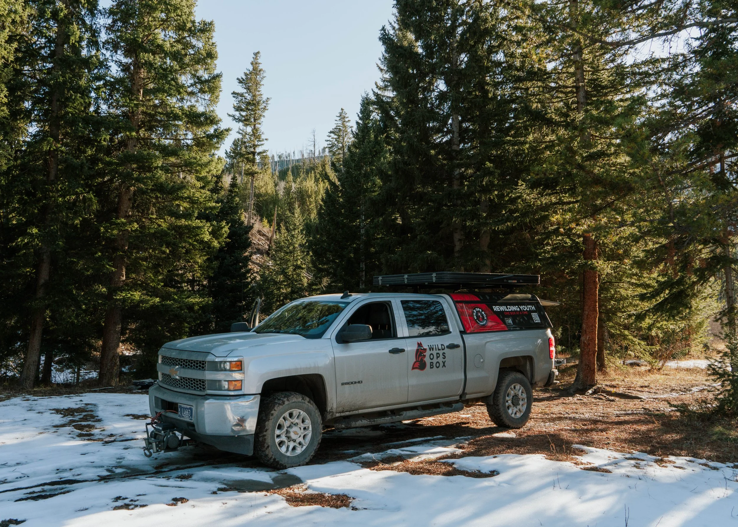 A silver Chevy Silverado pickup truck with a decal reading 'Wild Ops Box' on the door, parked on a dirt path with patches of snow, surrounded by tall evergreen trees in a forested area.