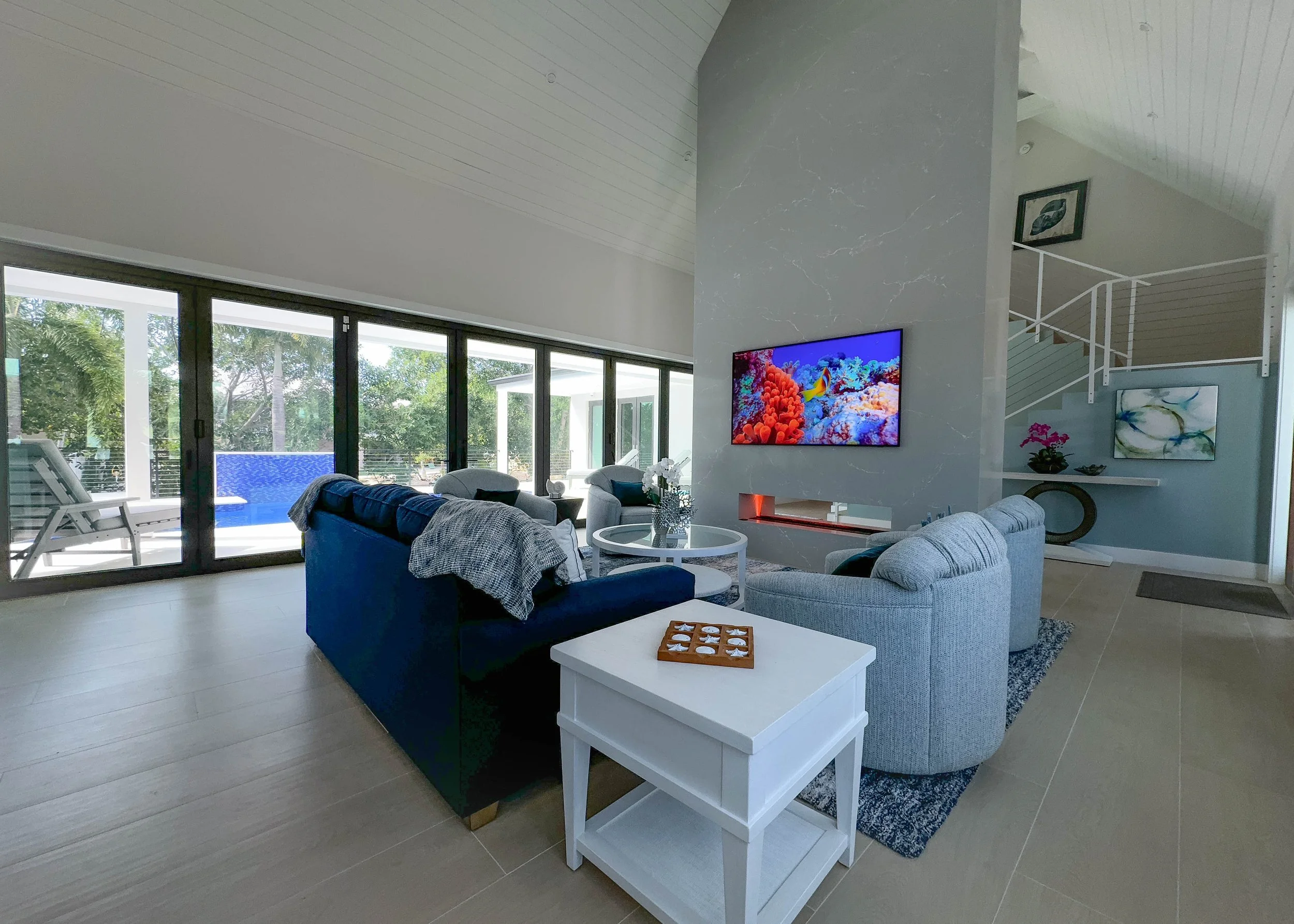 Living room with beige tile floor, light-colored walls, a large flat-screen TV on a modern fireplace, and seating area with a dark blue sofa, light gray ovoid chairs, and a white side table. Large glass sliding doors open to an outdoor balcony with a swimming pool and lounge chairs, overlooking trees.