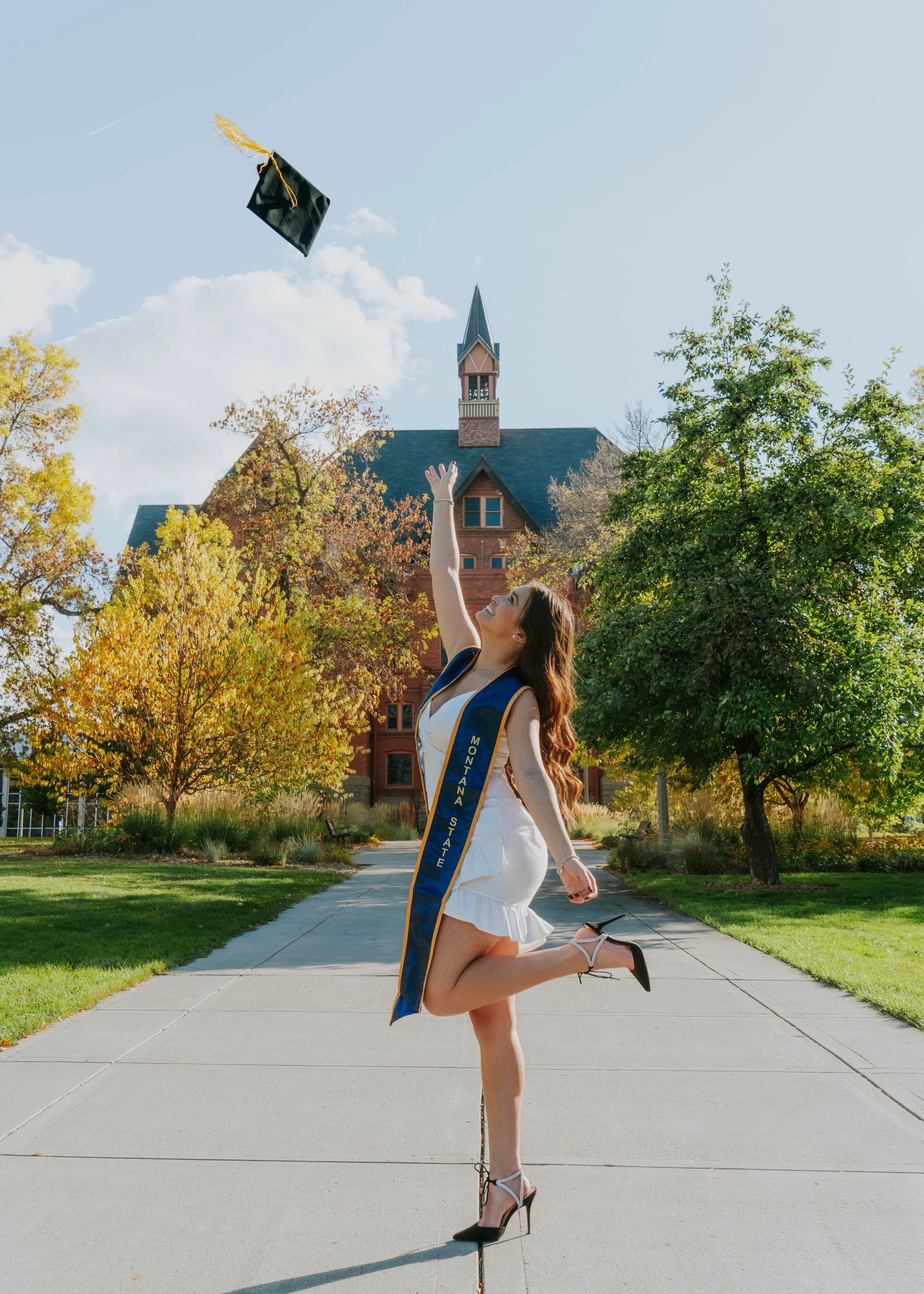 A woman in a white dress and black high heels is celebrating her graduation on a university campus, throwing her graduation cap into the air.