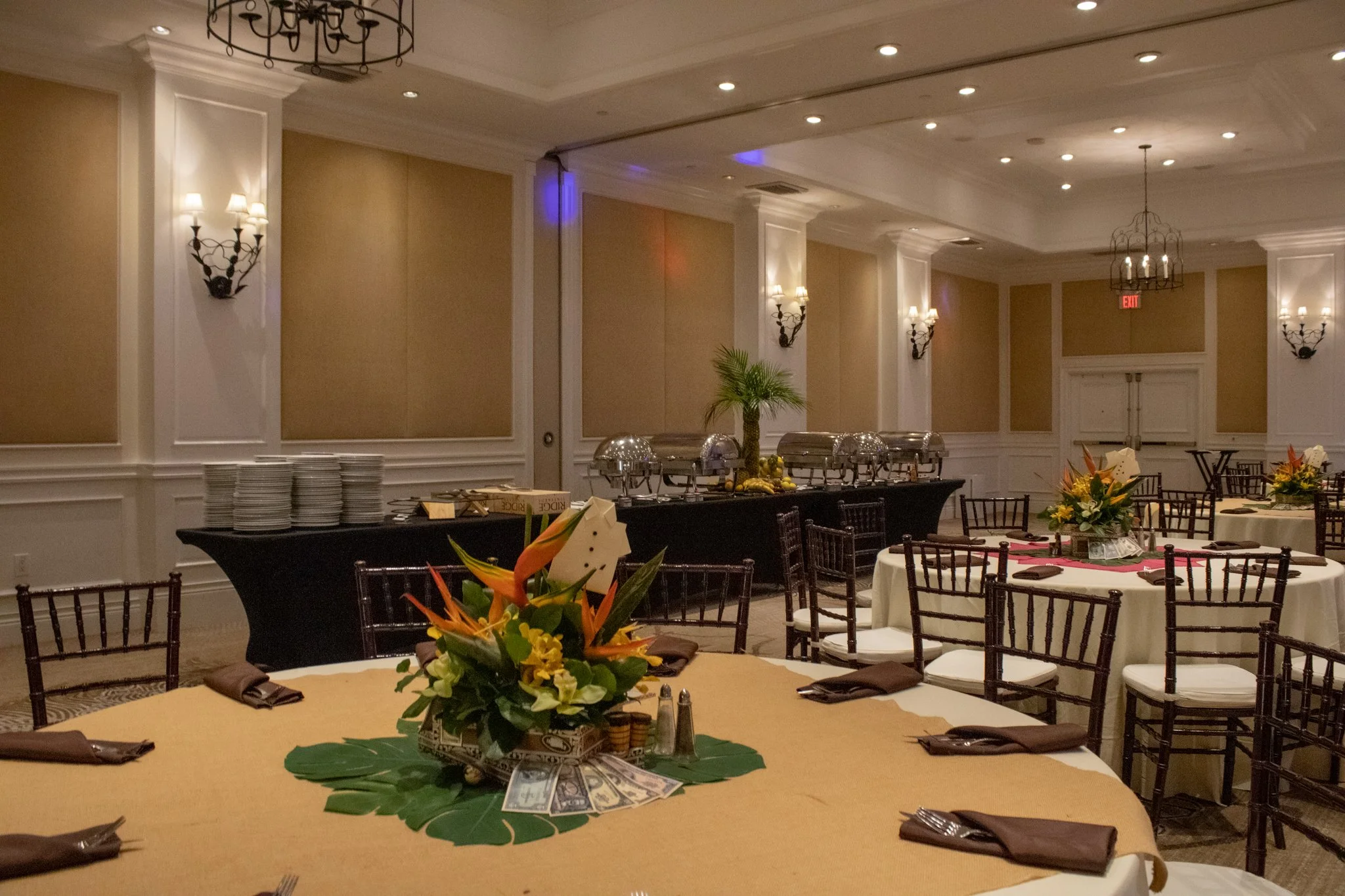 Banquet hall decorated for event with round tables covered in white and yellow tablecloths, floral centerpieces, and chairs with white cushions, set up alongside a buffet table with chafing dishes and plates, in a well-lit elegant room with wall sconces and chandeliers.