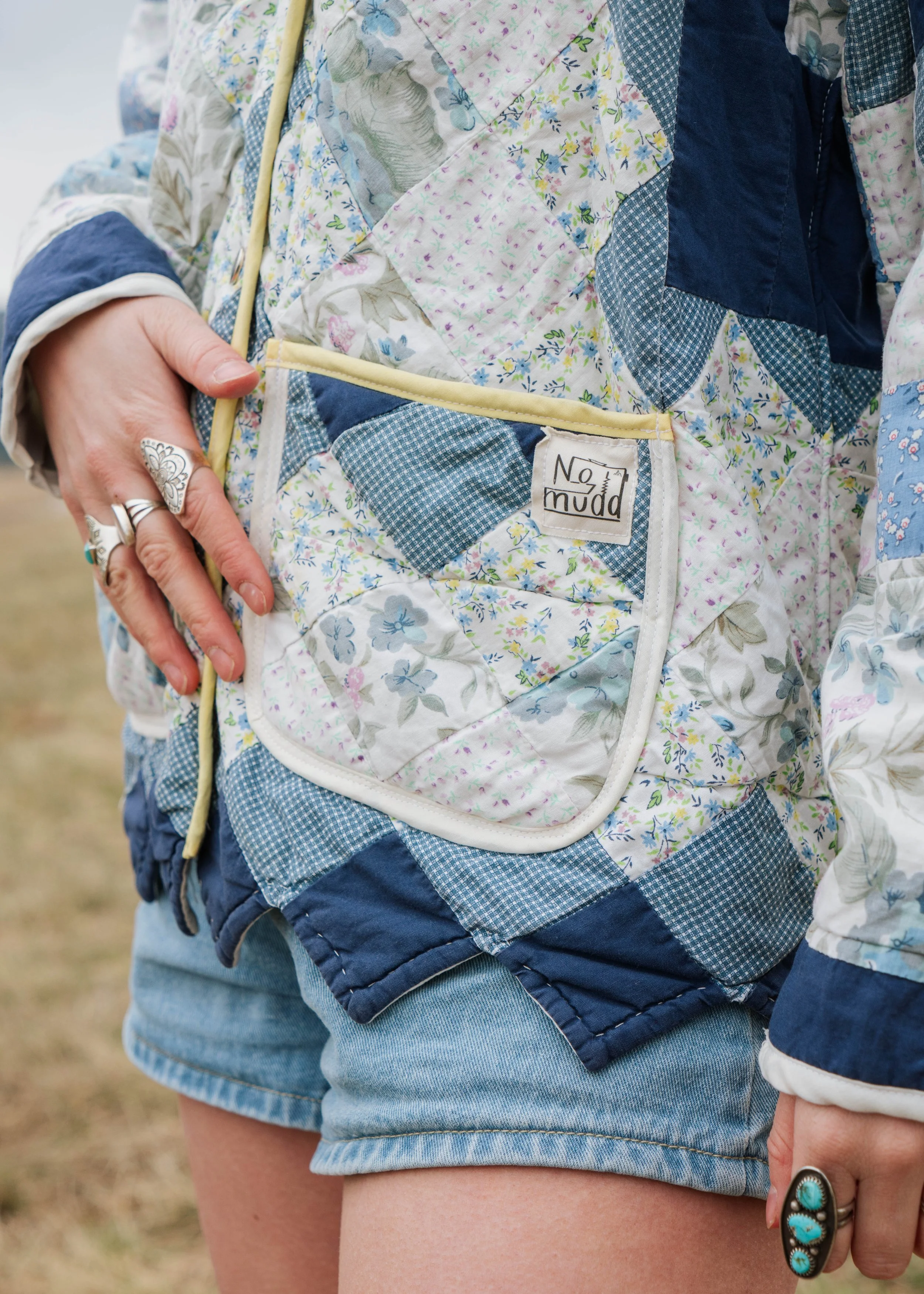 Close-up of a person wearing a patchwork quilted jacket, showing the pocket with a 'No Mudd' label, and layered rings on their fingers.