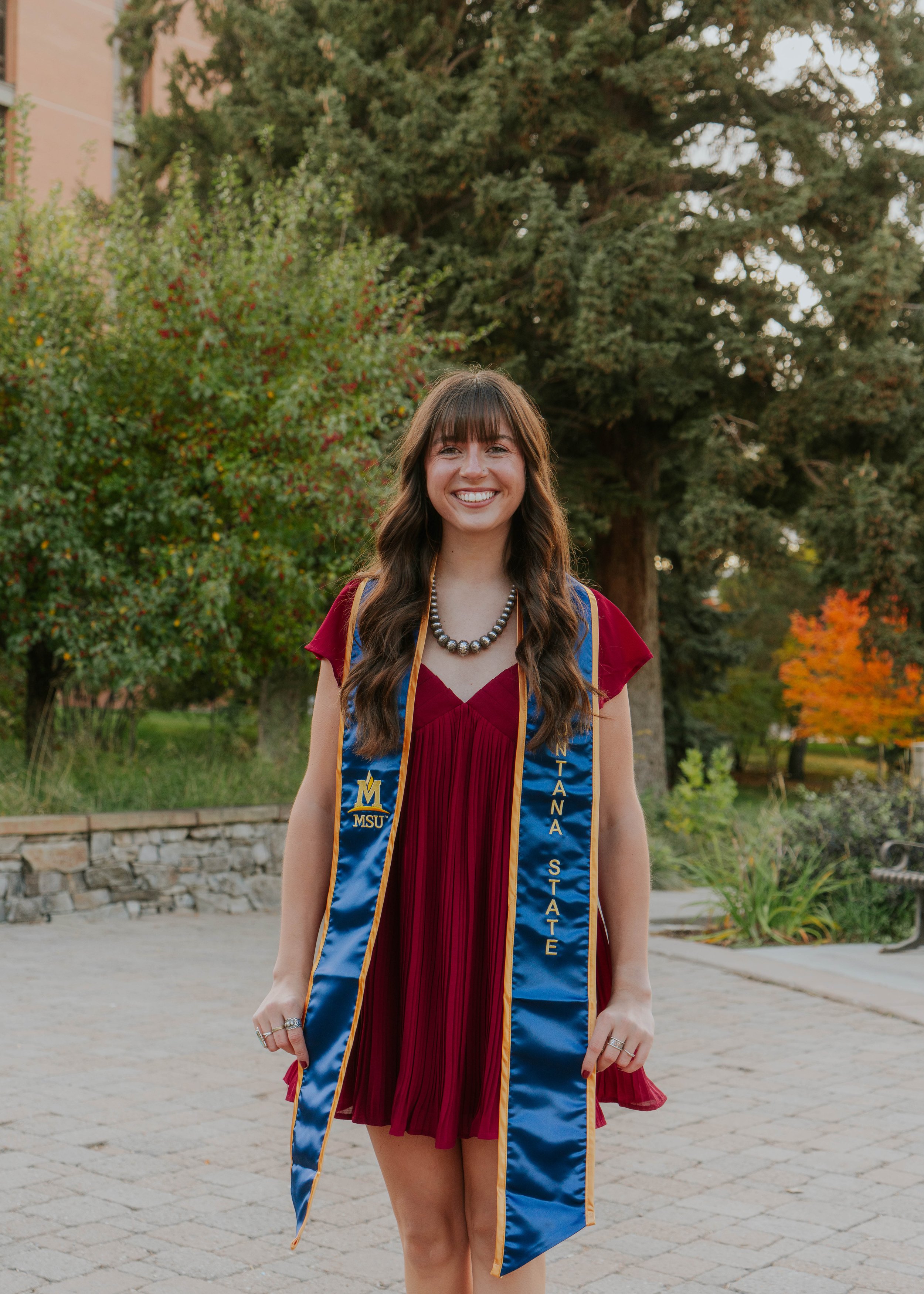 A young woman in a red dress and graduation gown with a blue and gold stole stands outdoors on a paved area, smiling. She is wearing a pearl necklace and rings, with trees and autumn foliage in the background.