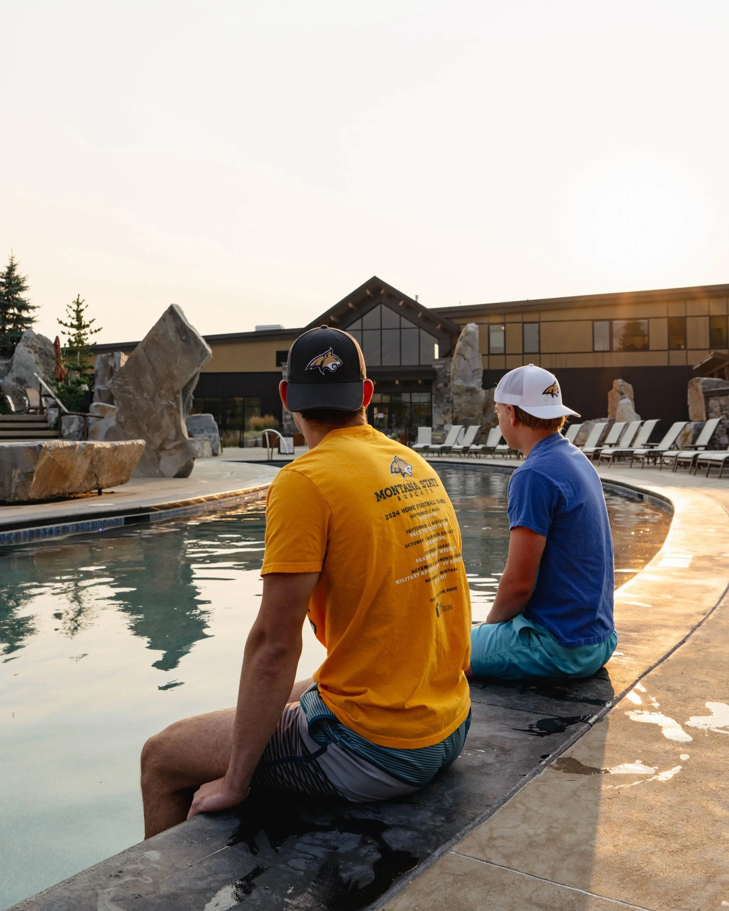 Two young men with sports caps sitting by a swimming pool during sunset, with a modern building and lounge chairs in the background.