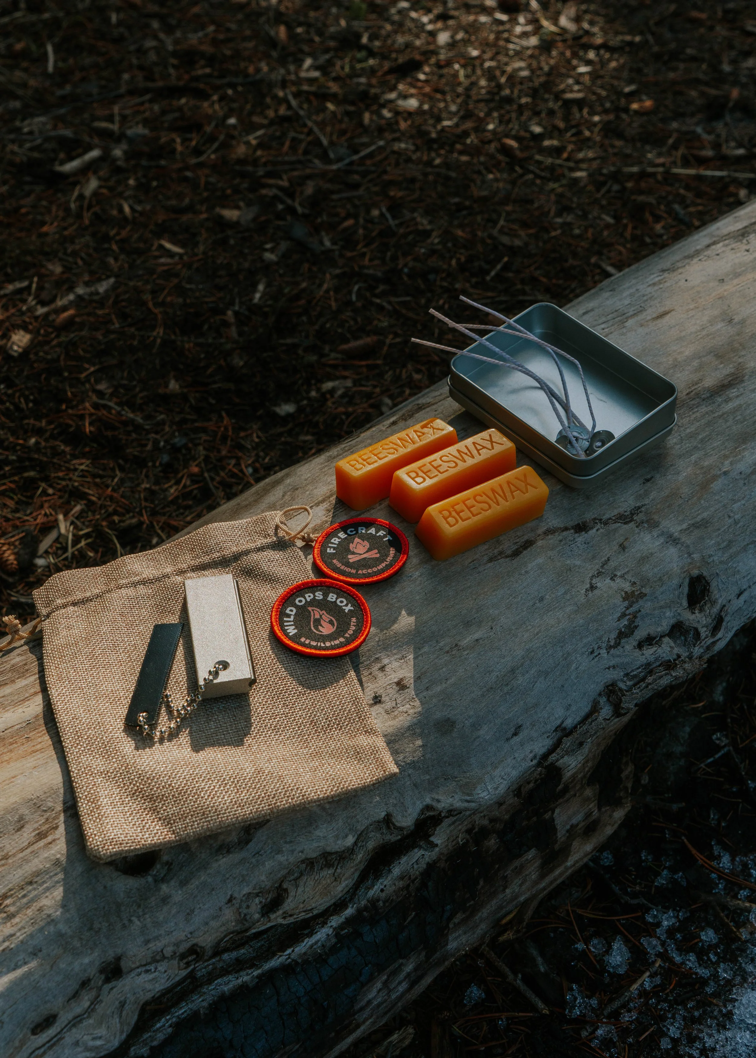 Camping gear and supplies arranged on a fallen log, including beeswax bars, patches that say 'fire craft' and 'wild ops box', a lighter, and a small metal box with lanyards.