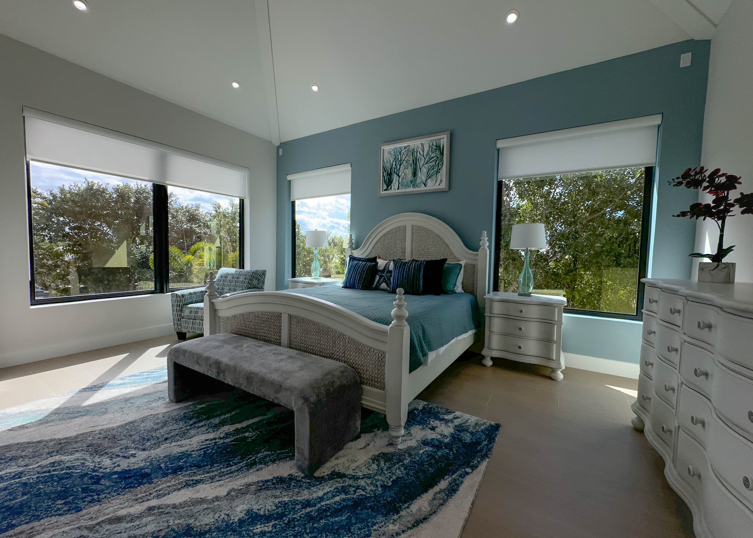 A bright bedroom with large windows showing a view of trees outside. The room has a white bed with a wicker headboard, decorated with dark pillows and a teal bedspread. There are two white nightstands with lamps, a gray bench at the foot of the bed, and a patterned rug on the wooden floor. A framed artwork hangs above the bed, and a white dresser with a potted plant on top is on the right side.