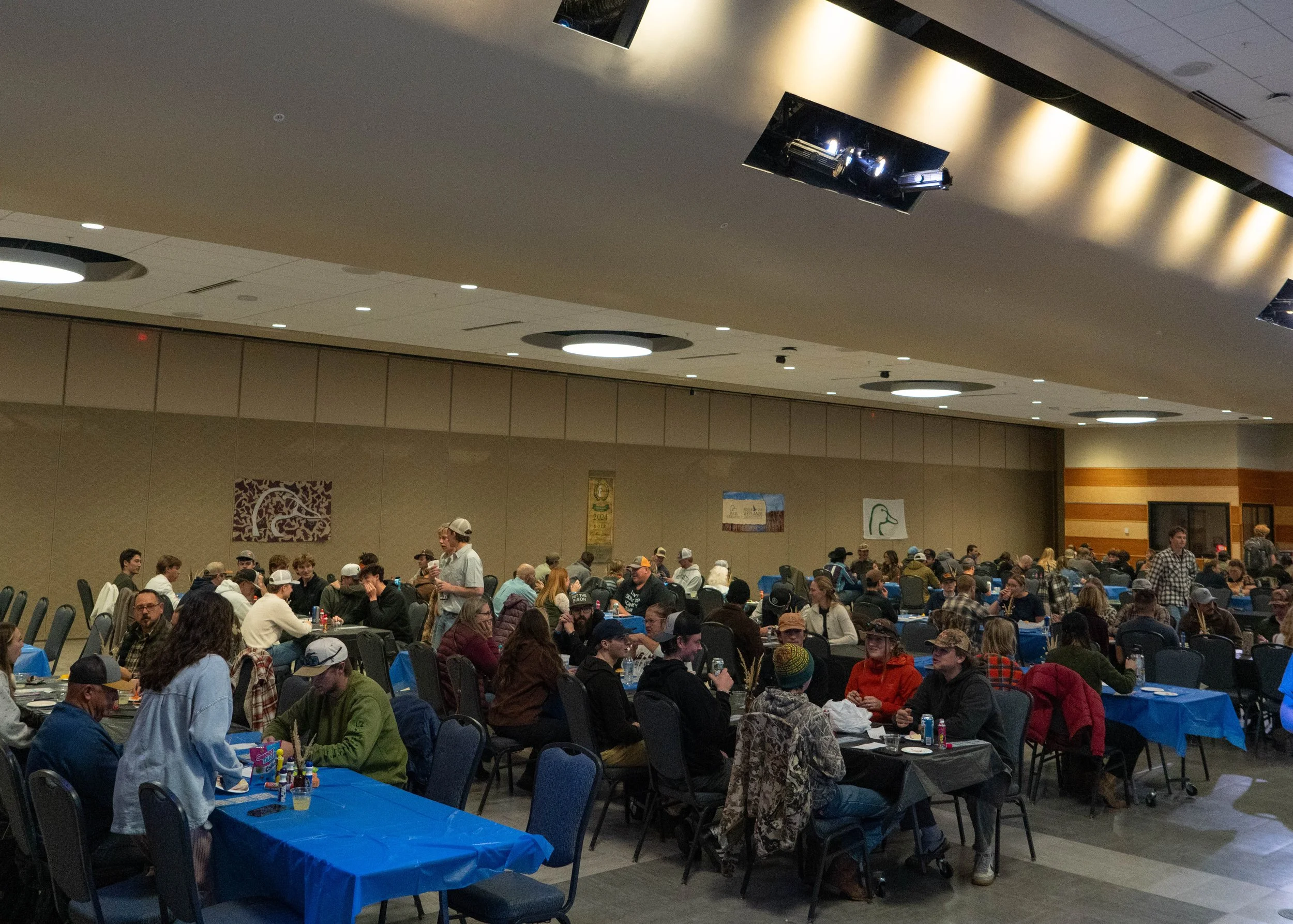A large indoor gathering with many people sitting at tables with blue tablecloths, some wearing hats, in a conference or event hall.