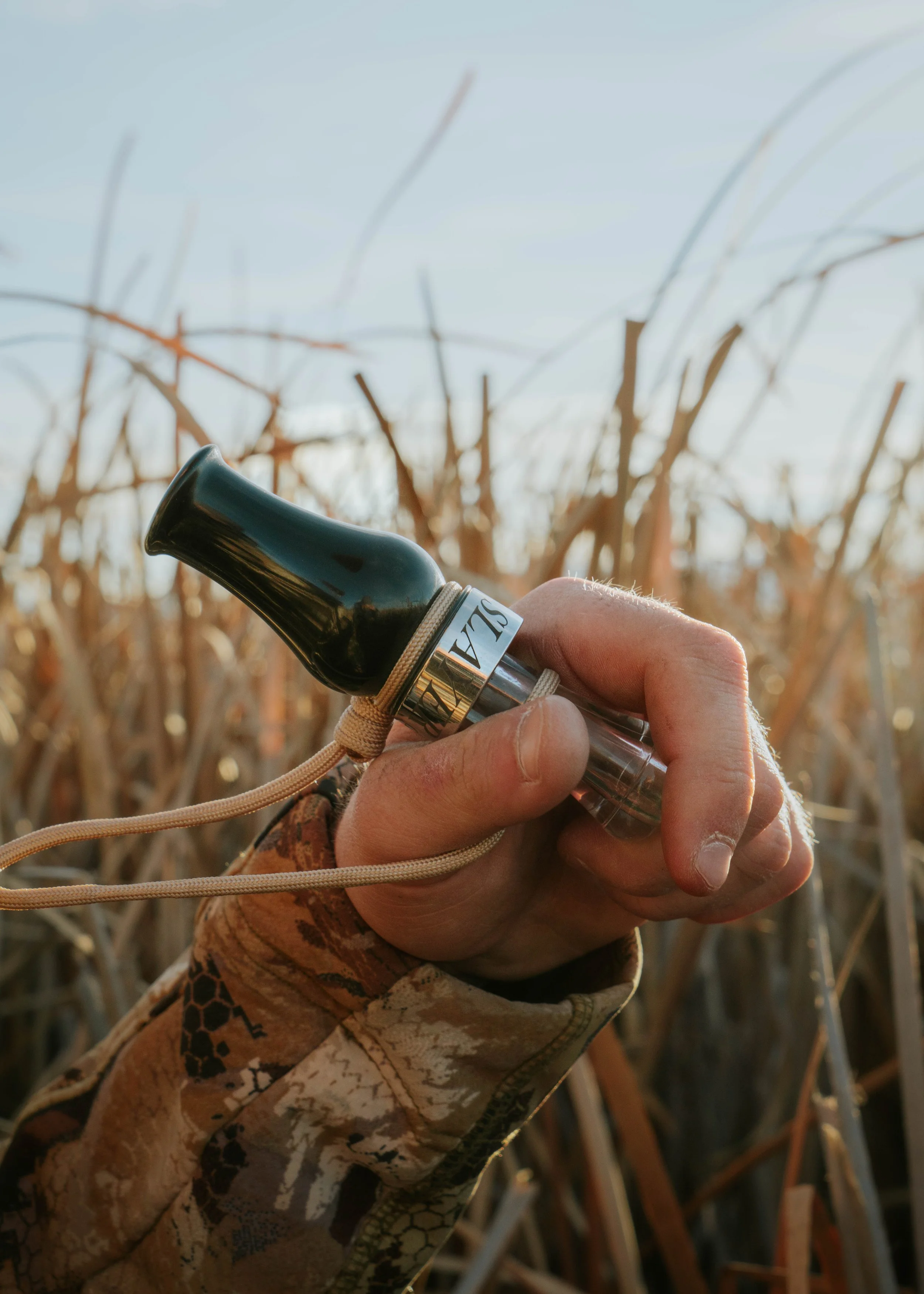 A hand holding a glass call used for bird watching or hunting, in a field of dry tall grass or wheat under clear sky.