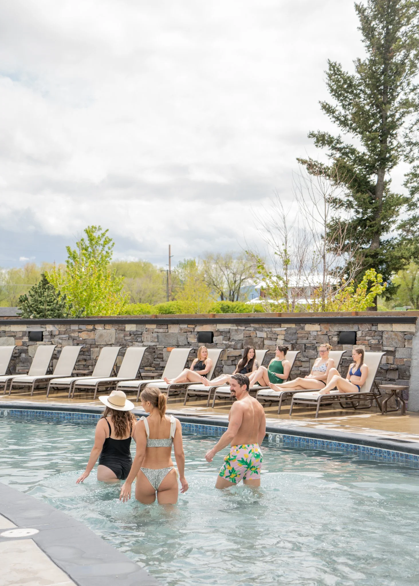 People relaxing by a poolside, some sitting on lounge chairs and others wading in the pool, with a stone wall and green trees in the background.