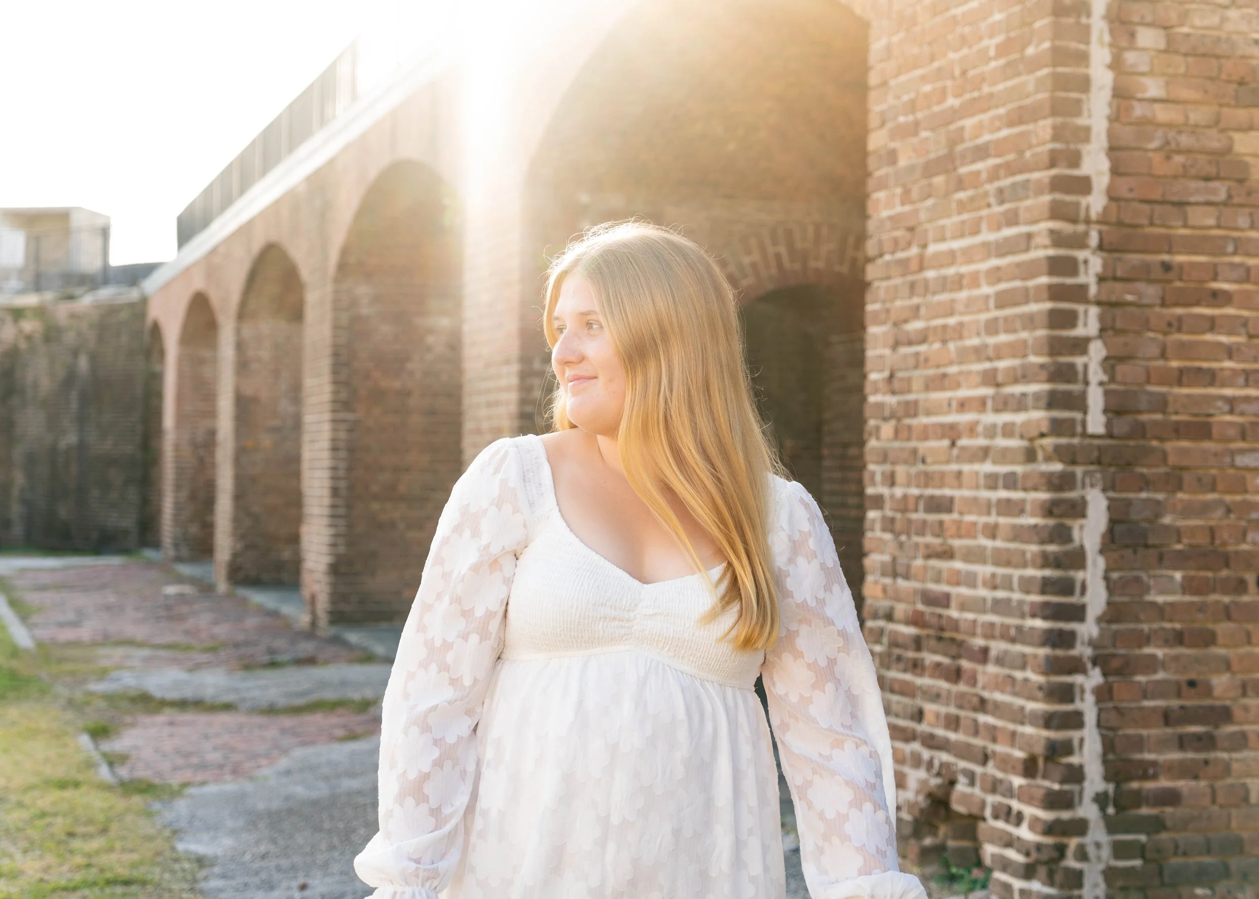 A young woman with long blonde hair wearing a white dress with sheer sleeves, standing outdoors near a brick wall with arches, backlit by the sun.