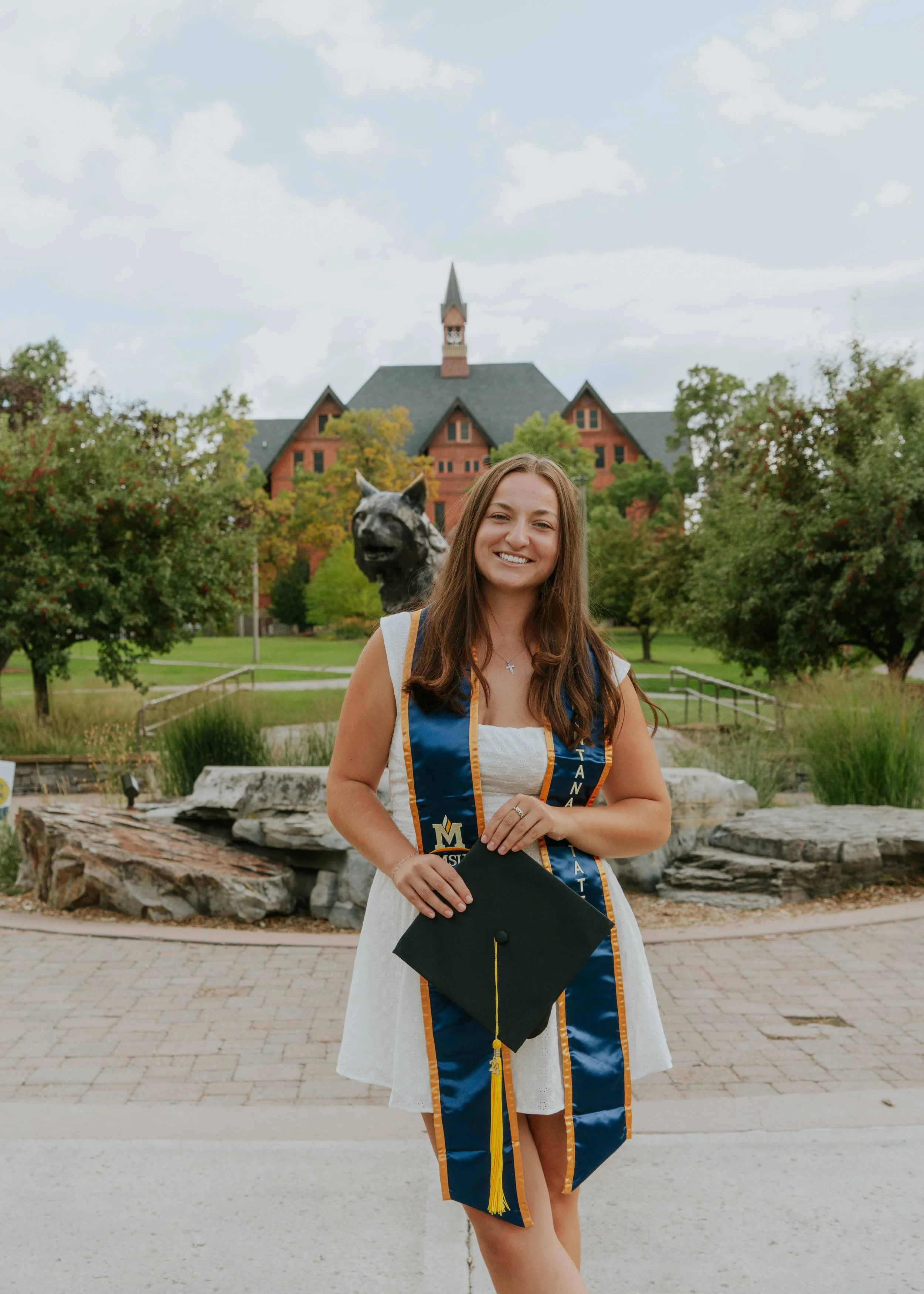 A young woman in a white dress and graduation sash holding a diploma cover, smiling outdoors on a graduation day, with a historic building and trees in the background.