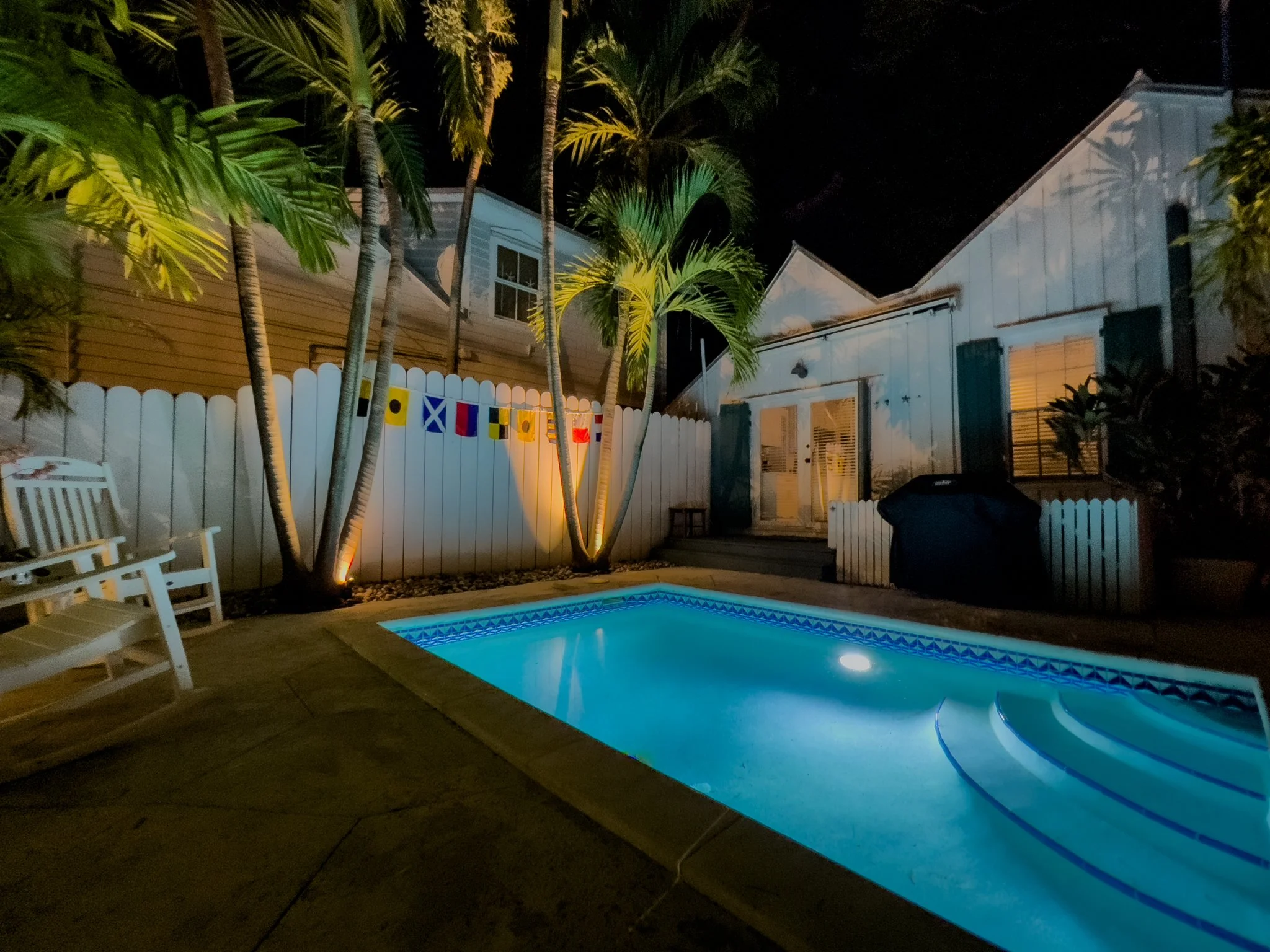 Nighttime backyard scene with a lit swimming pool, white wooden fence, and tropical palm trees. House with lit windows and deck visible in the background.