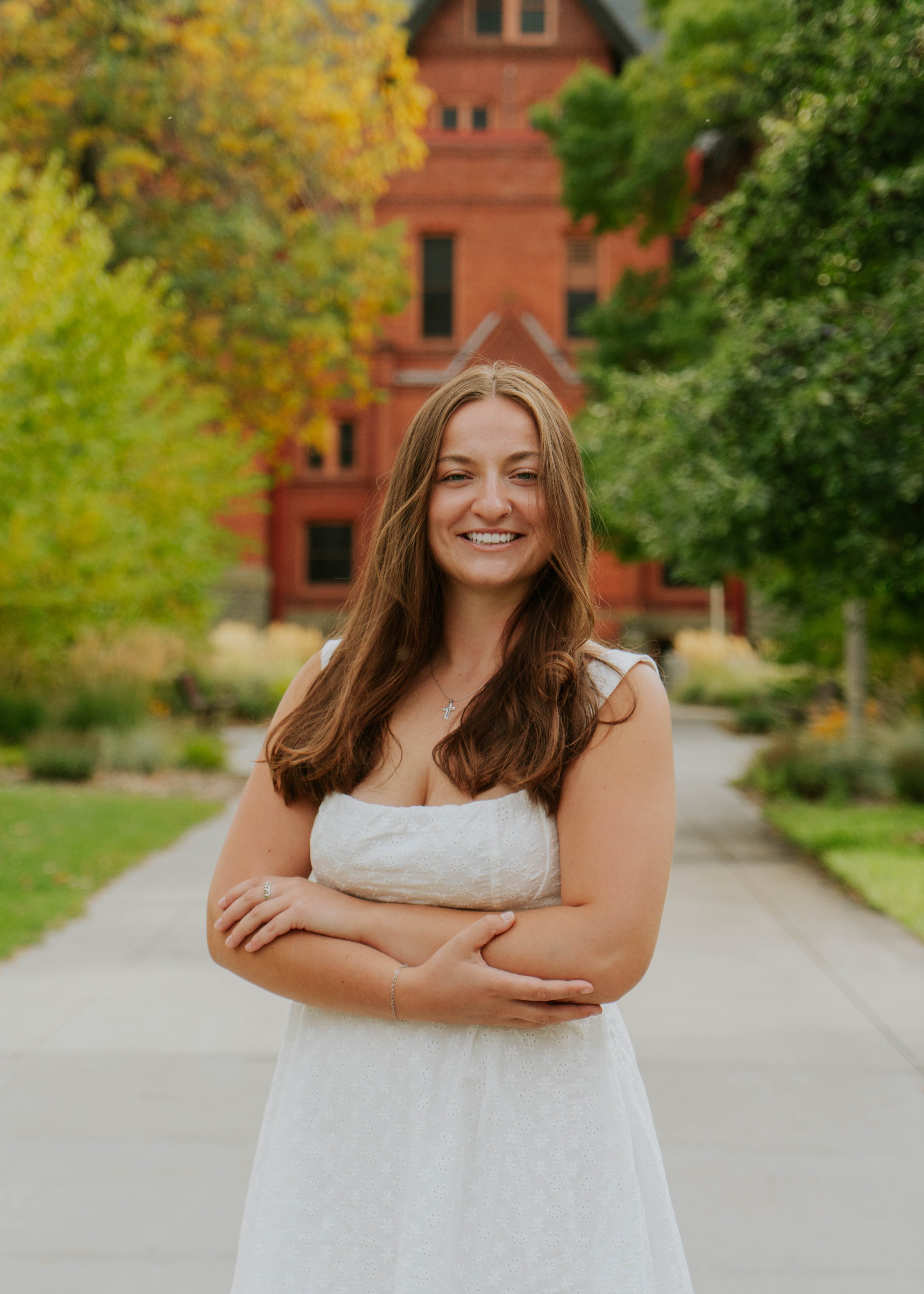 A young woman with brown hair and a white dress standing outdoors on a sidewalk with trees and a red brick building in the background.